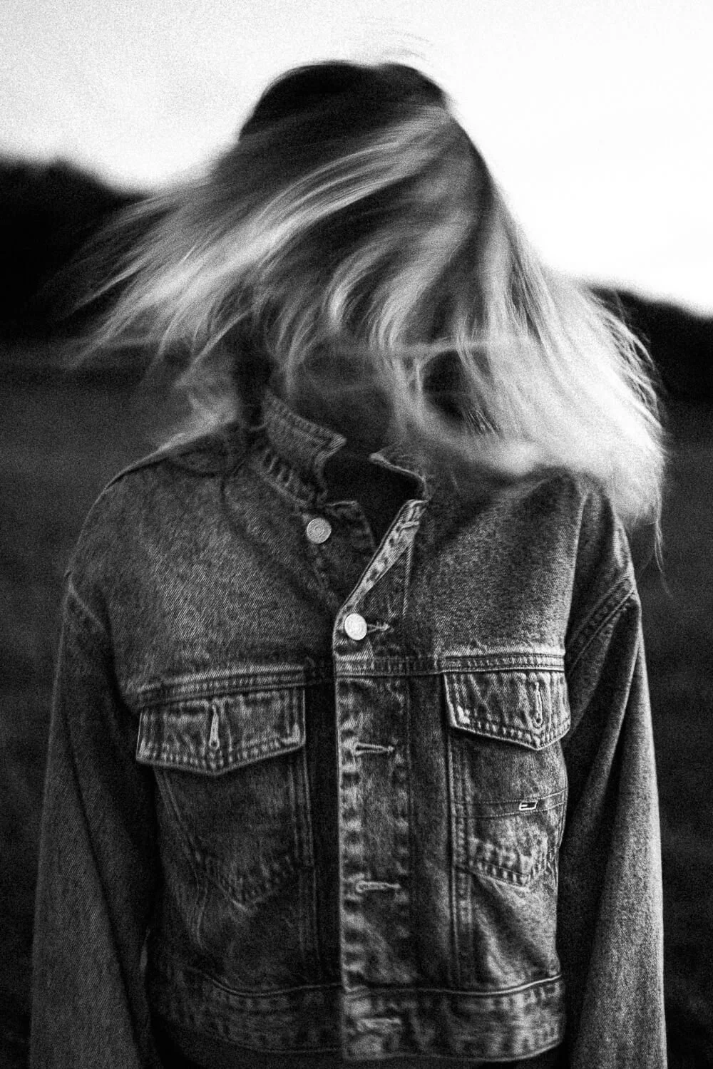 Black and white photo of a person with long hair covering their face, wearing a denim jacket.