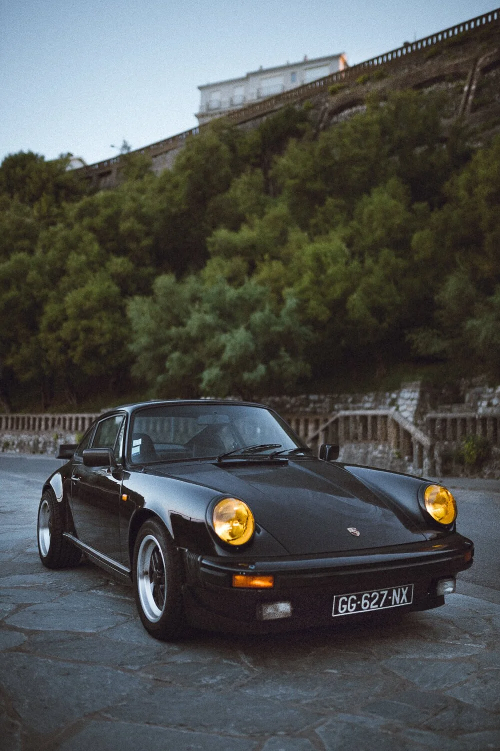 A black classic Porsche 911 sports car with yellow headlights parked on a stone-paved street at dusk, with green trees and a hillside with a building in the background.