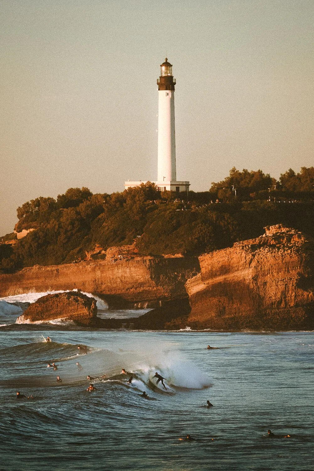 Surfers riding waves off a coast with a lighthouse atop a cliff in the background during sunset.