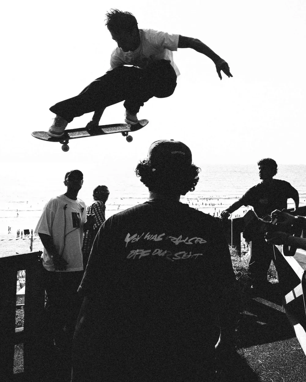 Un adolescent fait du skateboard en saut dans le ciel, vu en silhouette noire contre le ciel clair. D'autres personnes regardent ou attendent près de la plage en arrière-plan.