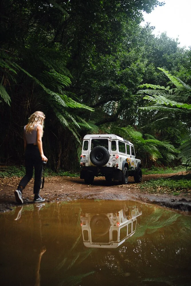 A woman walking near a muddy puddle on a forest trail with lush green foliage overhead and a white vintage off-road vehicle parked nearby, its reflection visible in the puddle.