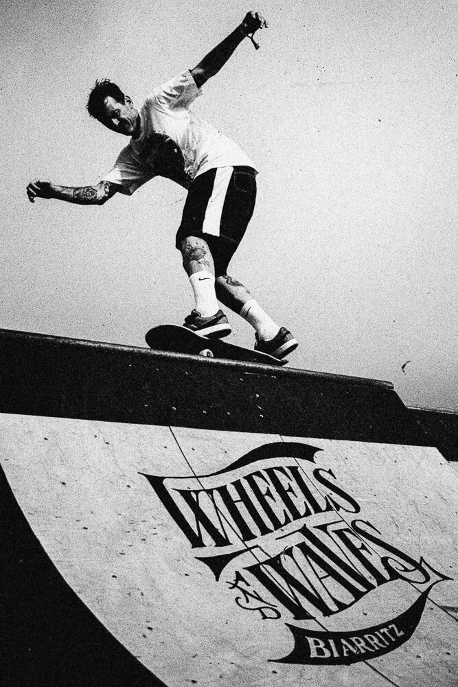 Skateboarder performing a trick on a ramp with 'Wheels and Waves Biarritz' written on it, black and white photograph.