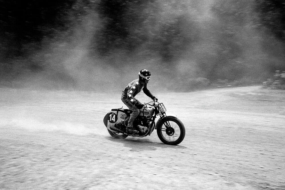 A person wearing a helmet riding a vintage motorcycle on a dirt track, kicking up dust.