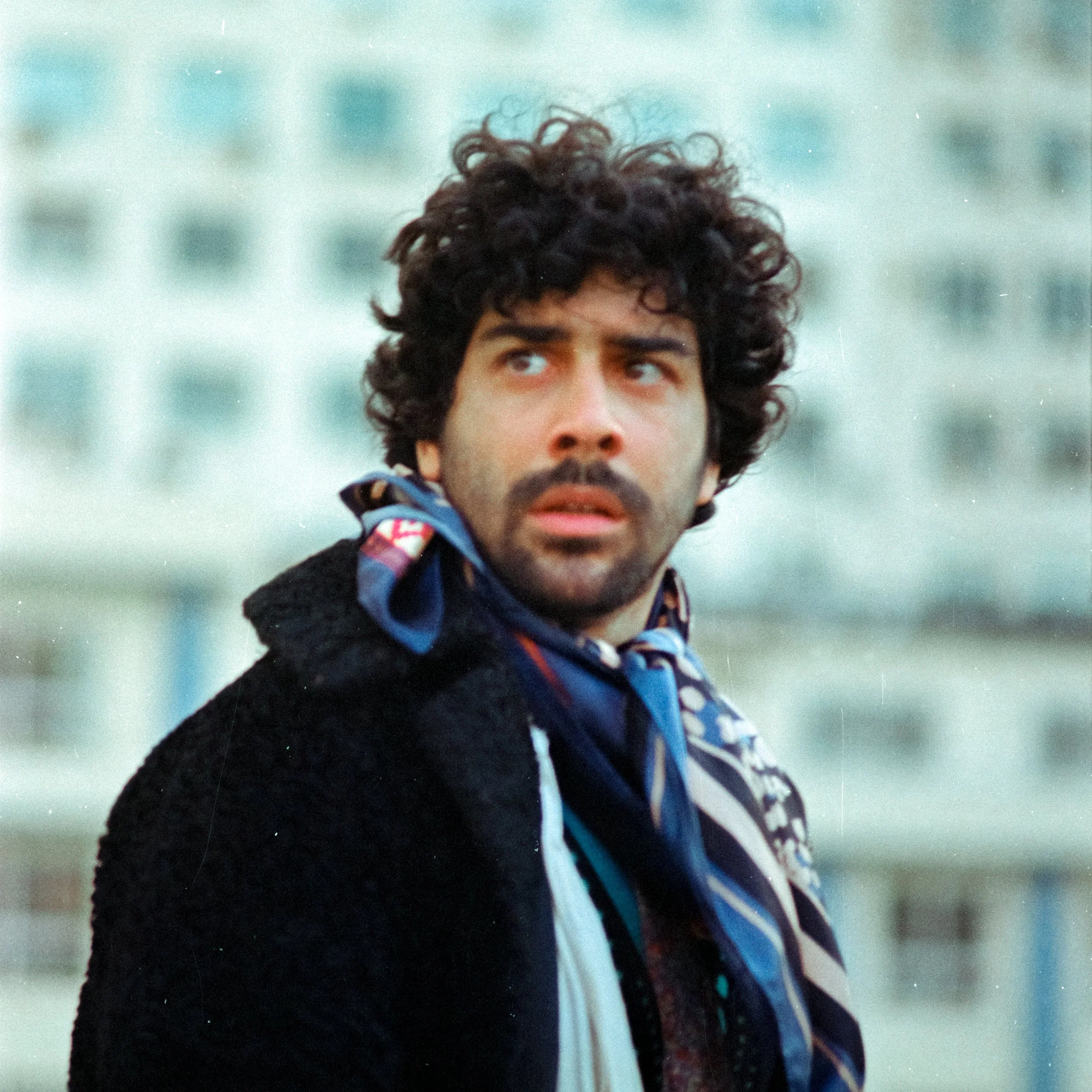 Homem com cabelo cacheado, barba e roupa escura olhando para o lado. Fundo desfocado com tonalidades de azul claro.