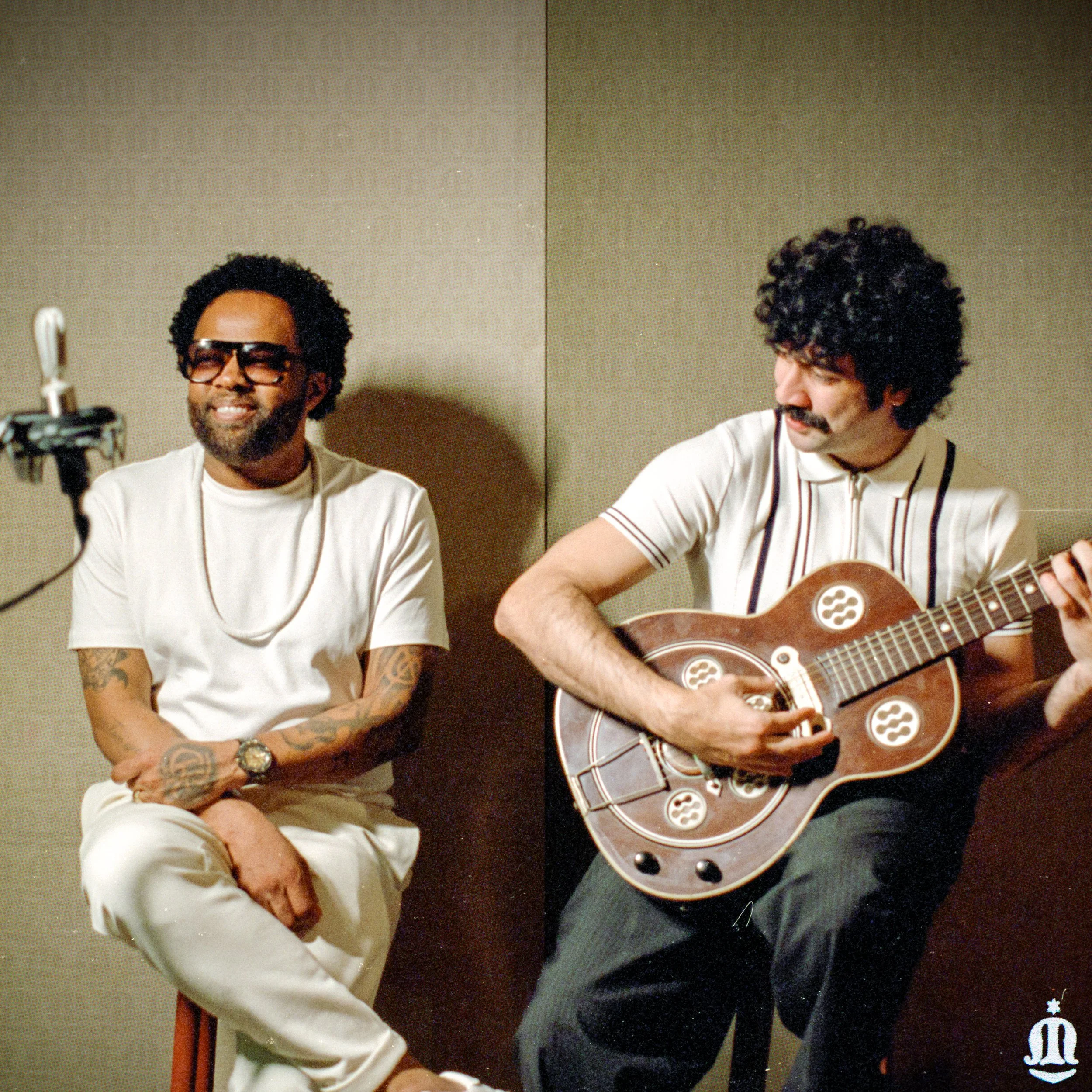 Dois homens jovens, um tocando violão, sentados em uma sala com fundo neutro.