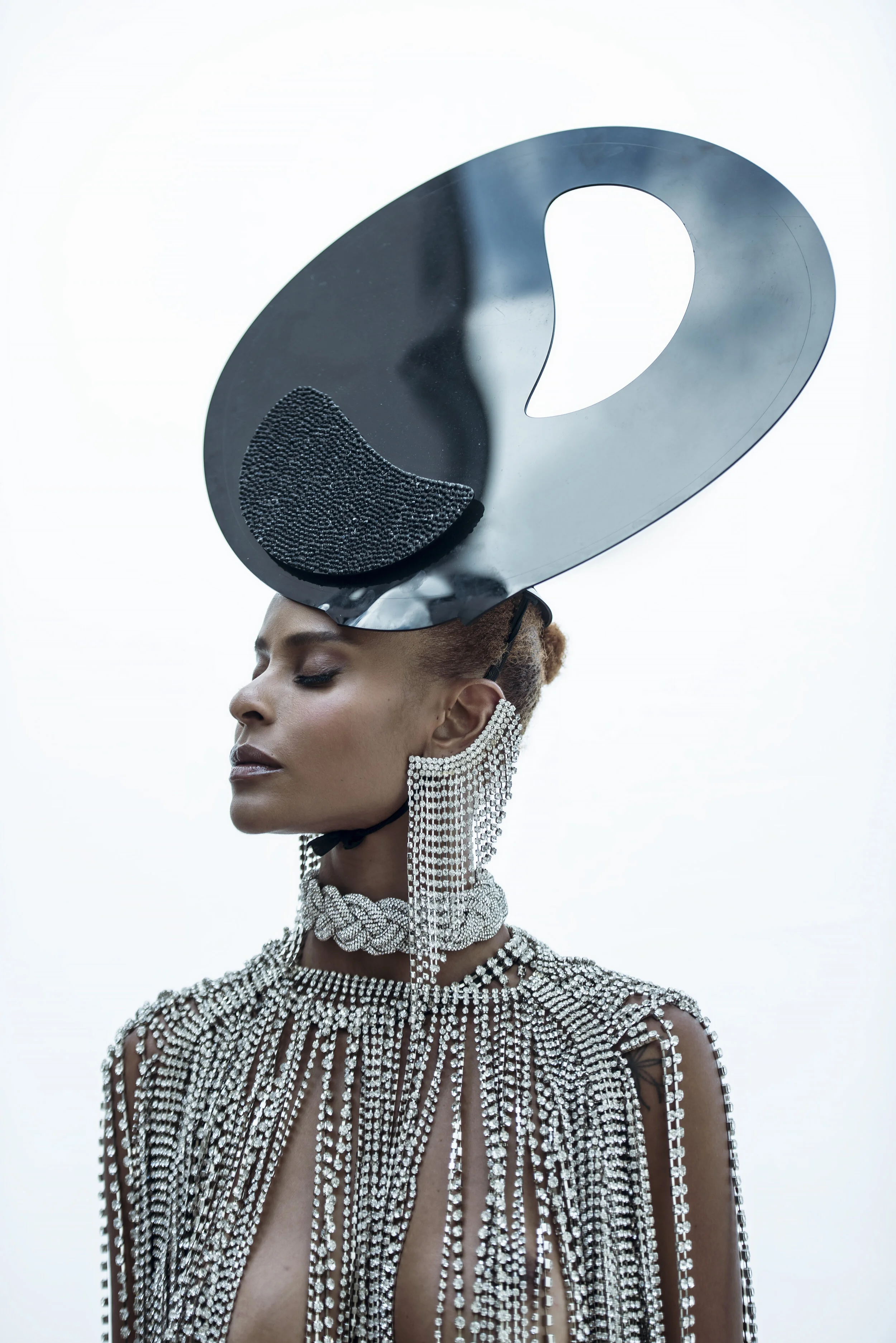 Mulher estilizada com acessórios de pérolas, usando um chapéu decorativo grande de metal com detalhes em preto, fechada com cabelos presos e com maquiagem sutil.