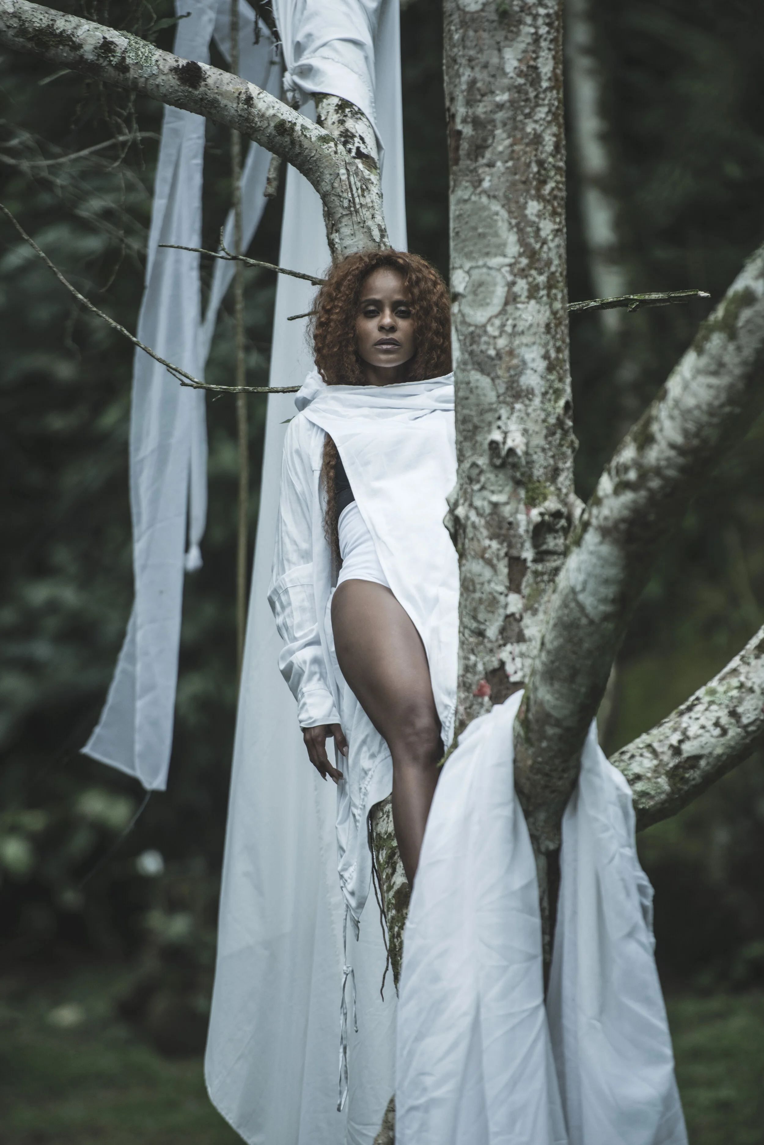 Mulher com cabelo cacheado e pele escura, vestindo roupa branca, com uma expressão séria, está encostada em uma árvore em uma floresta, com panos brancos pendurados ao seu redor.
