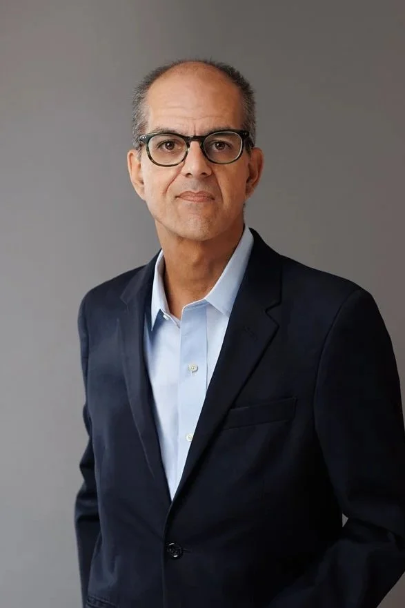 A middle-aged man with short hair, wearing glasses, a light blue dress shirt, and a dark suit jacket, standing against a gray background.