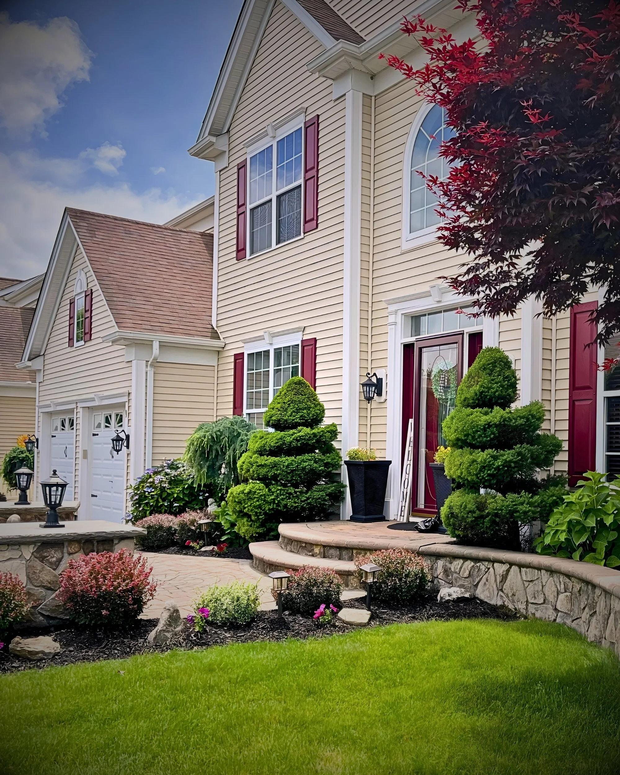 Front yard of a beige two-story suburban house with red shutters, a stone pathway, well-manicured lawn, and landscaped garden with shrubs, flowers, and small trees.