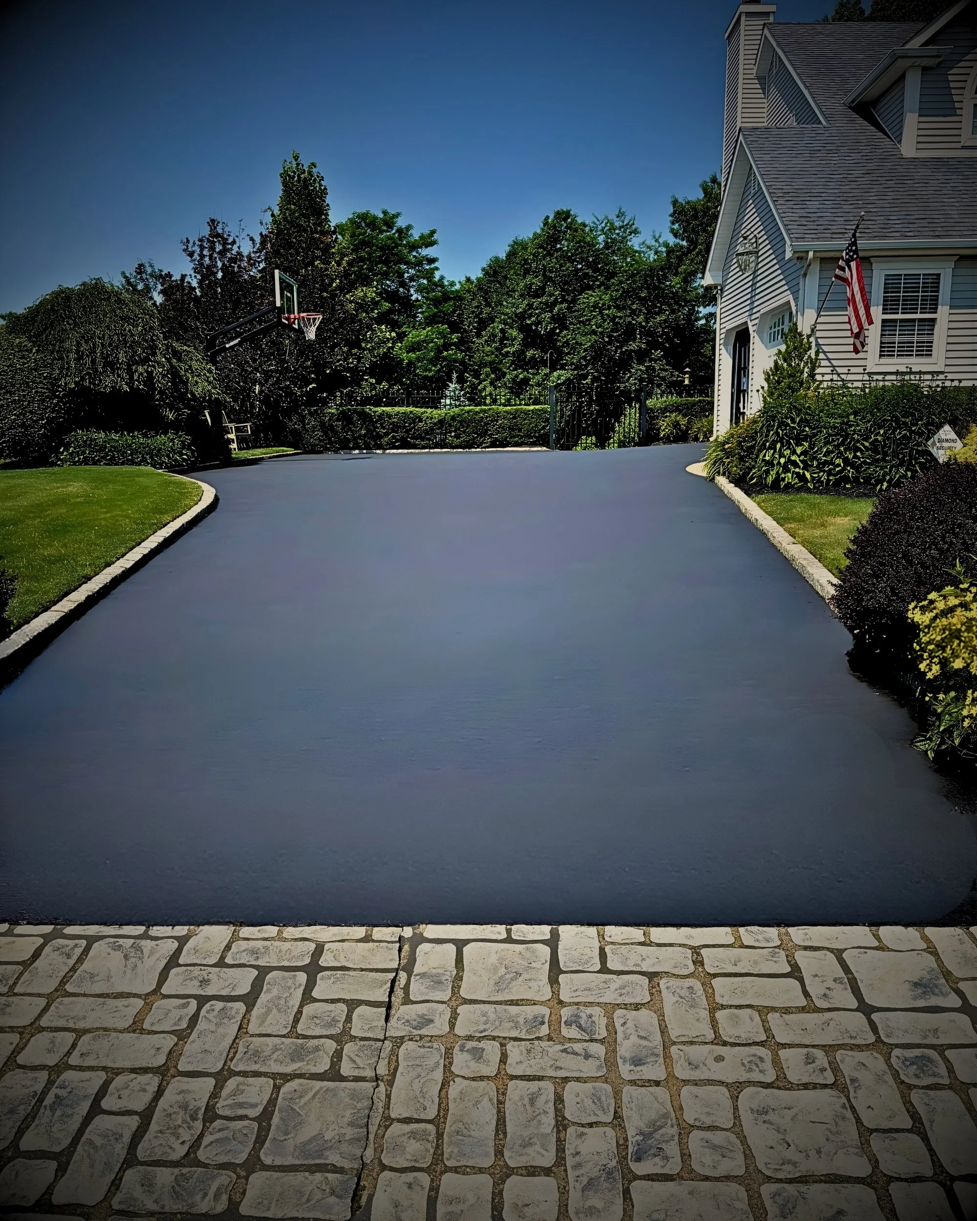 A driveway with fresh black asphalt leading to a large house, bordered by neatly trimmed grass and landscaped bushes, with a basketball hoop and lush green trees in the background under a clear blue sky.