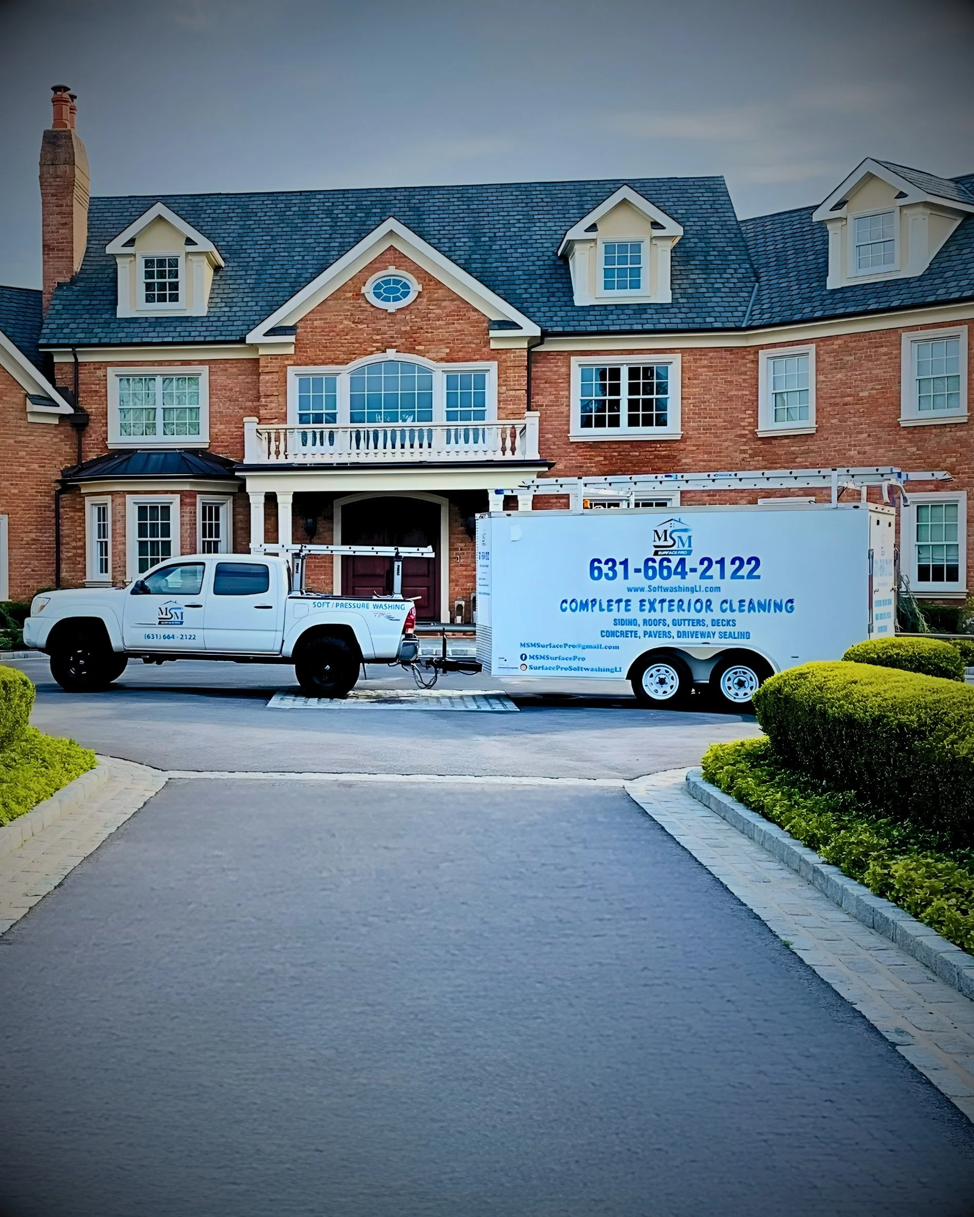 A large brick house with multiple windows and a balcony, a driveway with a truck and trailer for exterior cleaning services, and well-kept green shrubs along the sides.