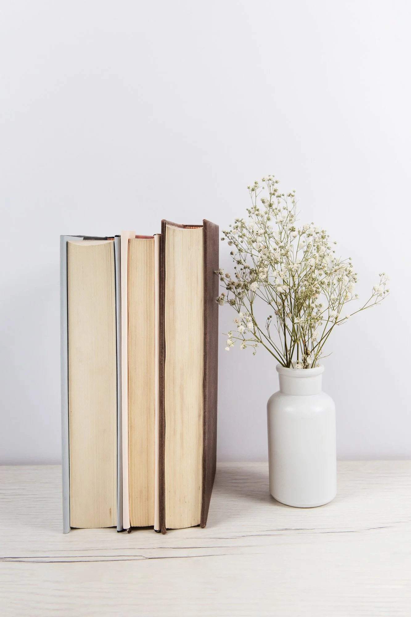 Five books standing upright next to a white vase with delicate white flowers on a light wooden surface.