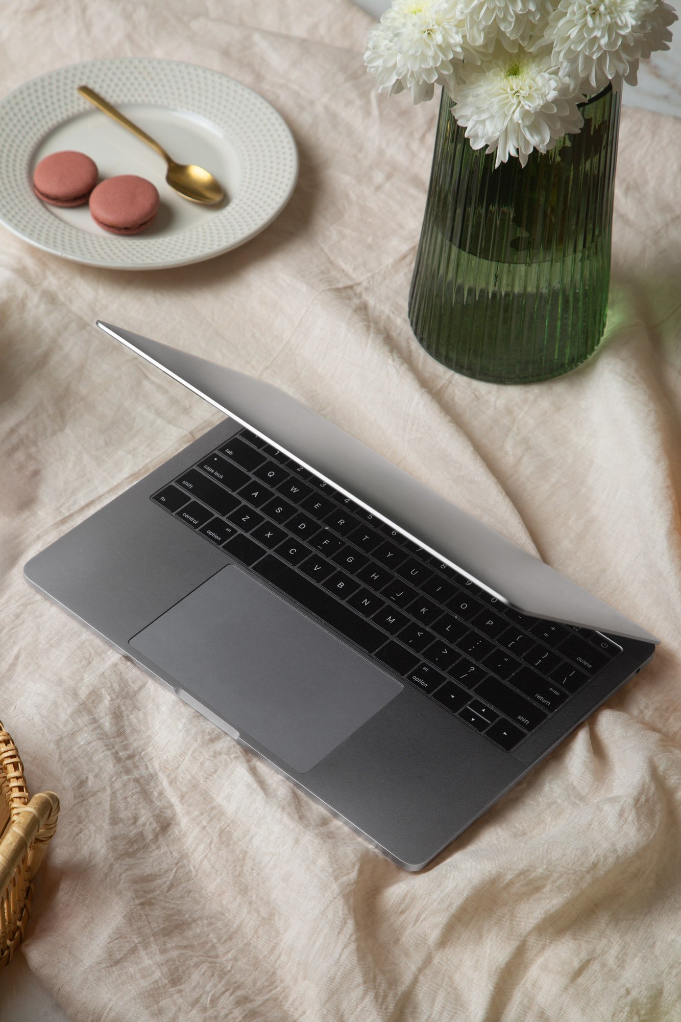 A close-up of a laptop with a black keyboard, a white trackpad, and a silver body placed on a beige fabric surface. In the background, there is a green, ribbed glass vase filled with white flowers and a white plate with two pink macarons and a gold spoon on a matching white saucer.