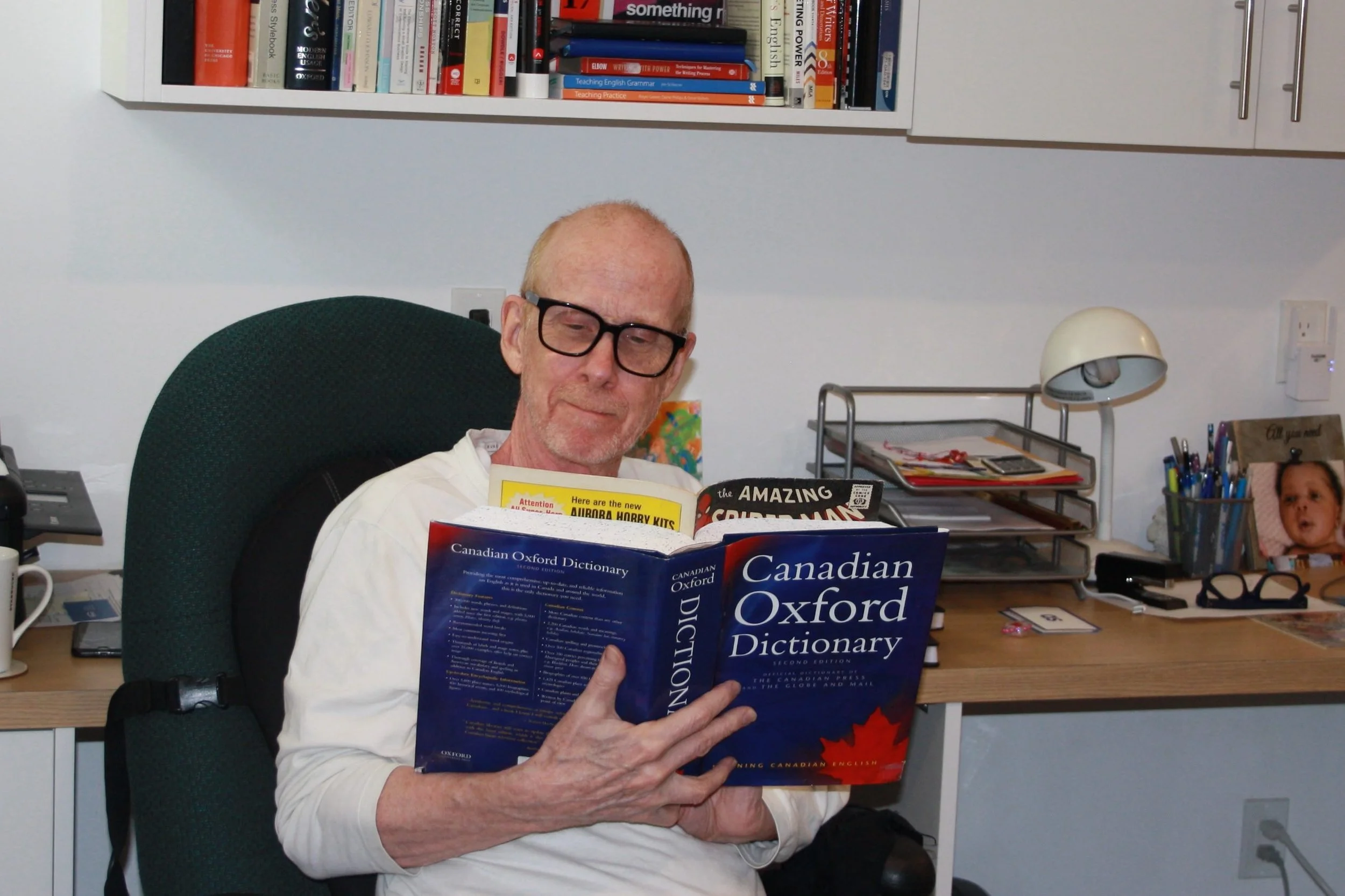 Keith Goddard is seated in his office, apparently reading a dictionary, though the edge a comic can been seen poking out from the dictionary.