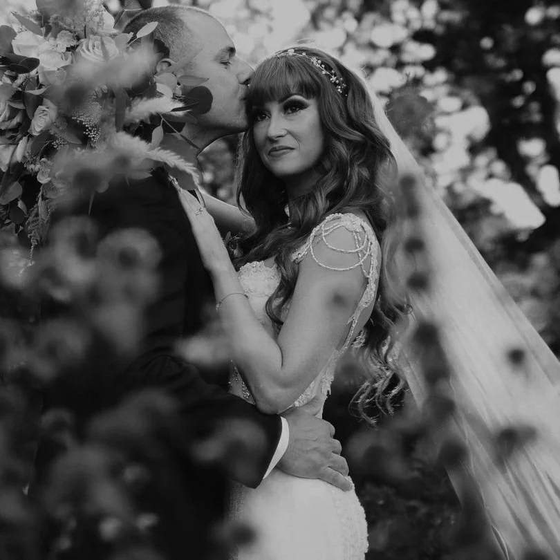 A black and white photo of a bride and groom outdoors, with the groom kissing the bride's forehead and the bride looking at the camera. The bride has long, wavy hair and wears a wedding dress with decorative shoulder details, while the groom is in a suit. Flowers and greenery surround them.