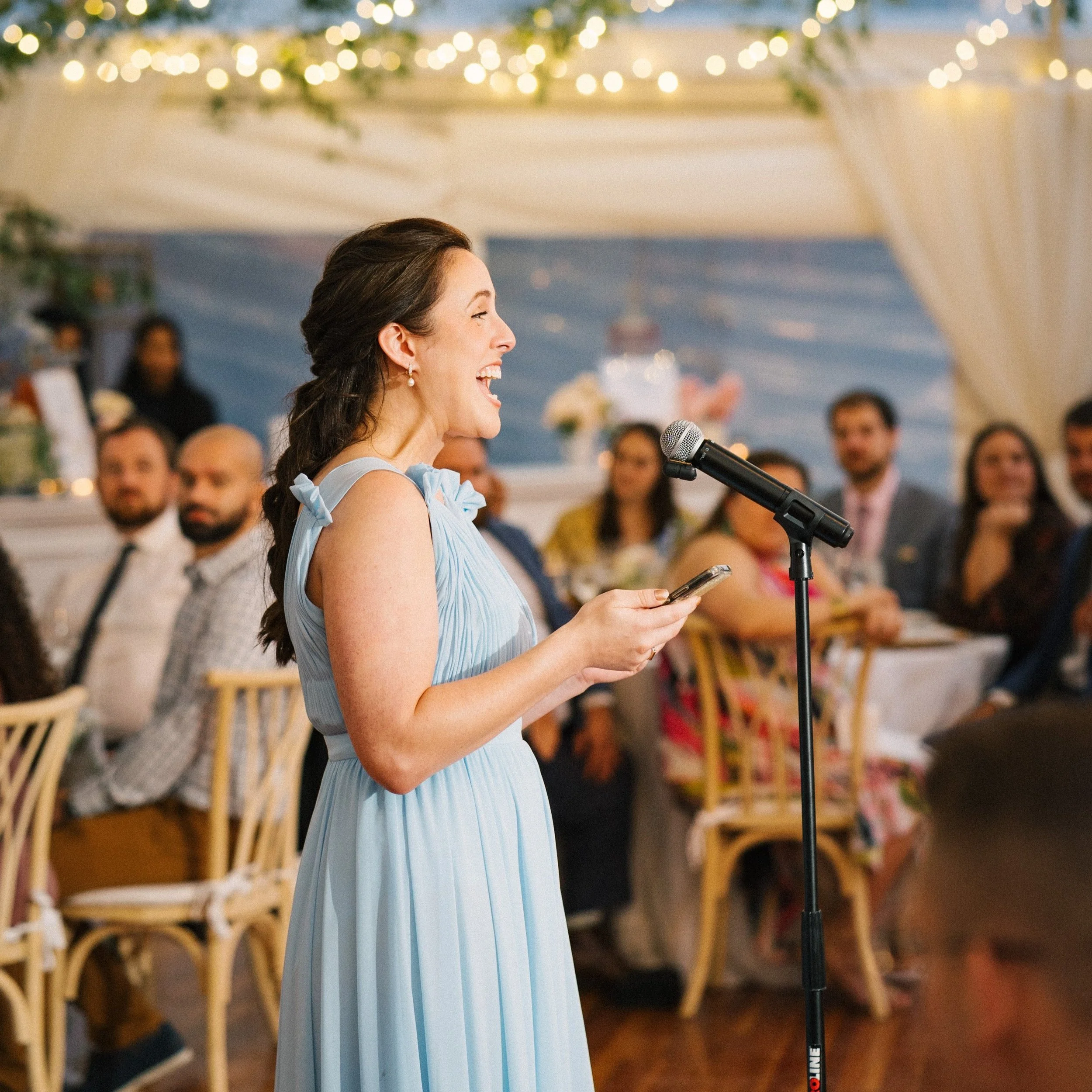 A woman in a light blue dress giving a speech at a decorated event with guests seated at tables in the background.