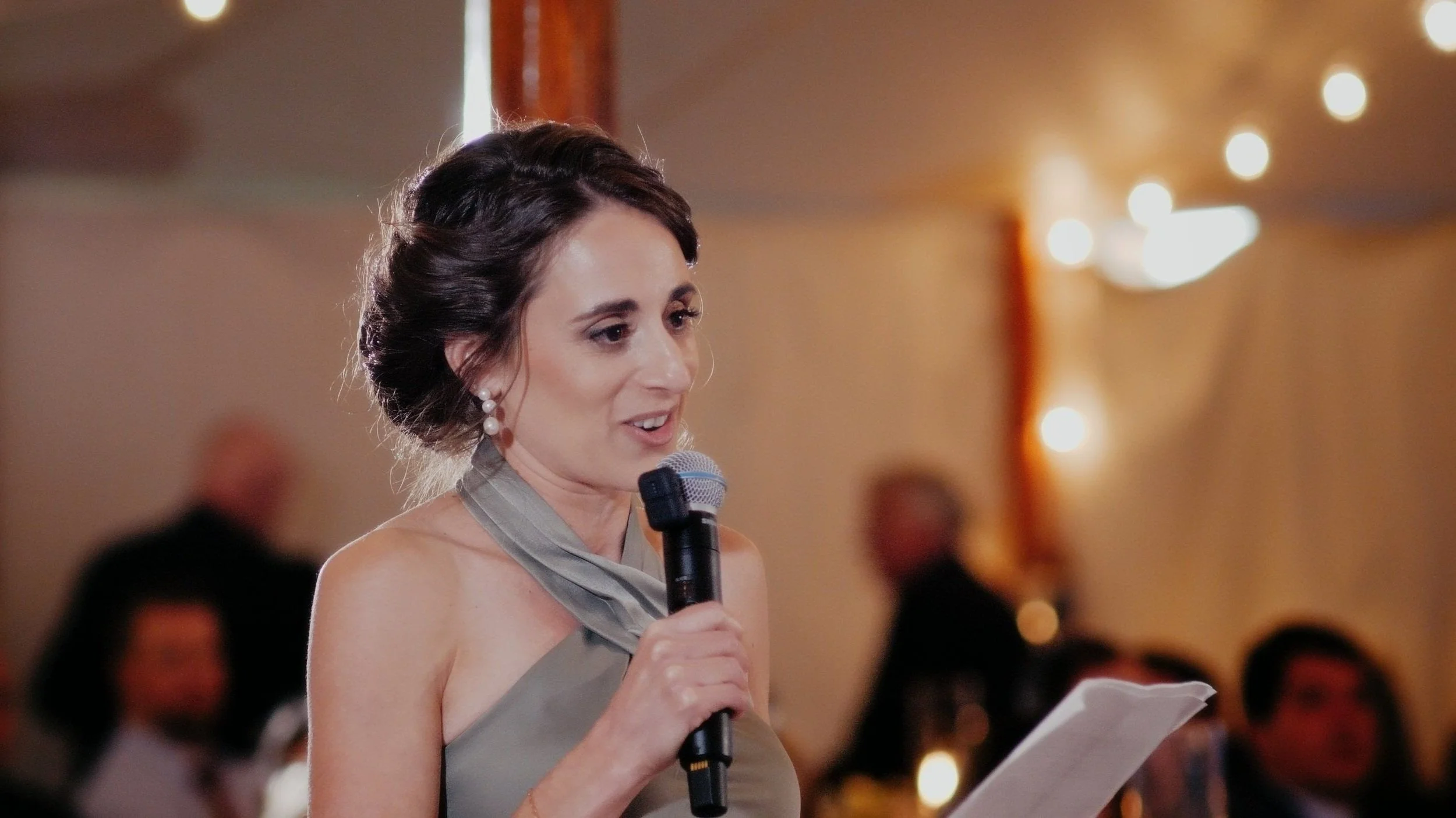 A woman with dark hair in an elegant updo, wearing pearl earrings and a gray dress with a matching scarf, speaking into a microphone and holding a paper at a formal event.