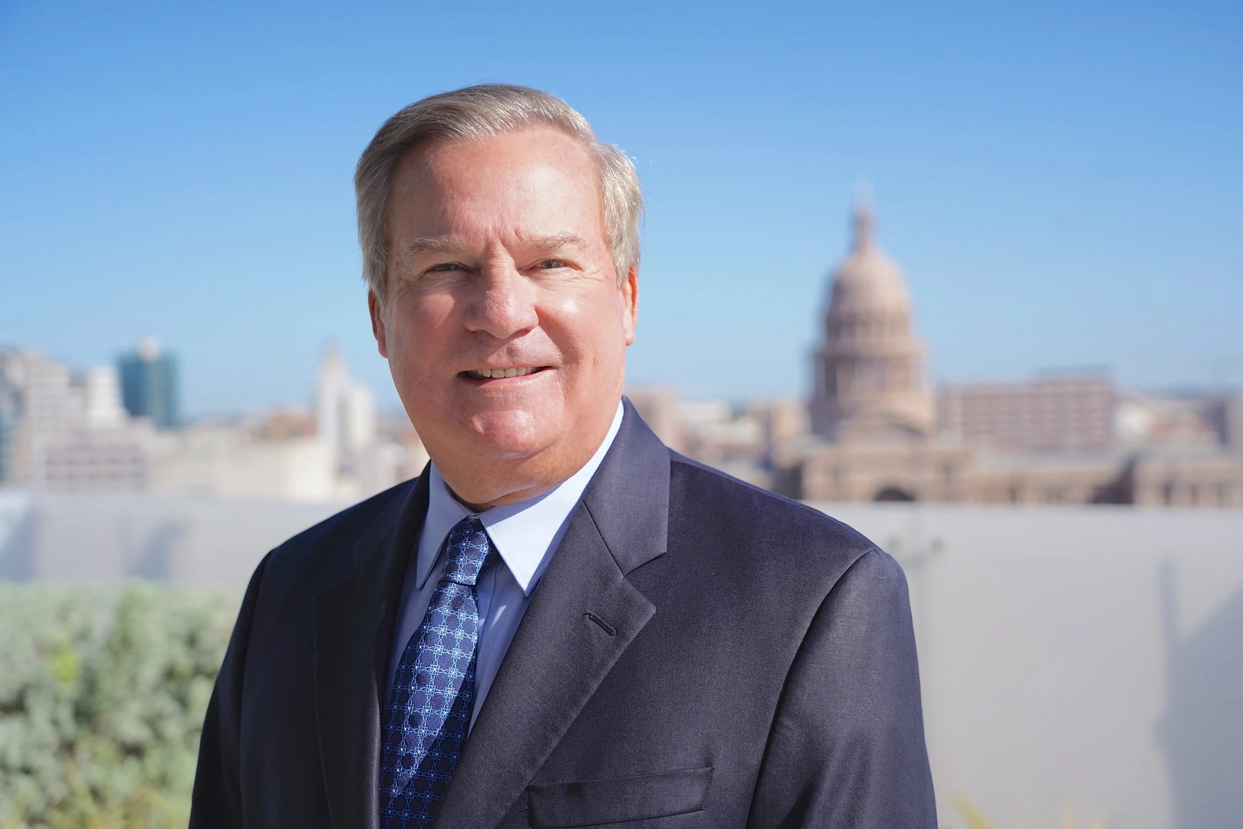 A man in a suit standing outdoors with a city skyline and Capitol building in the background on a clear day.