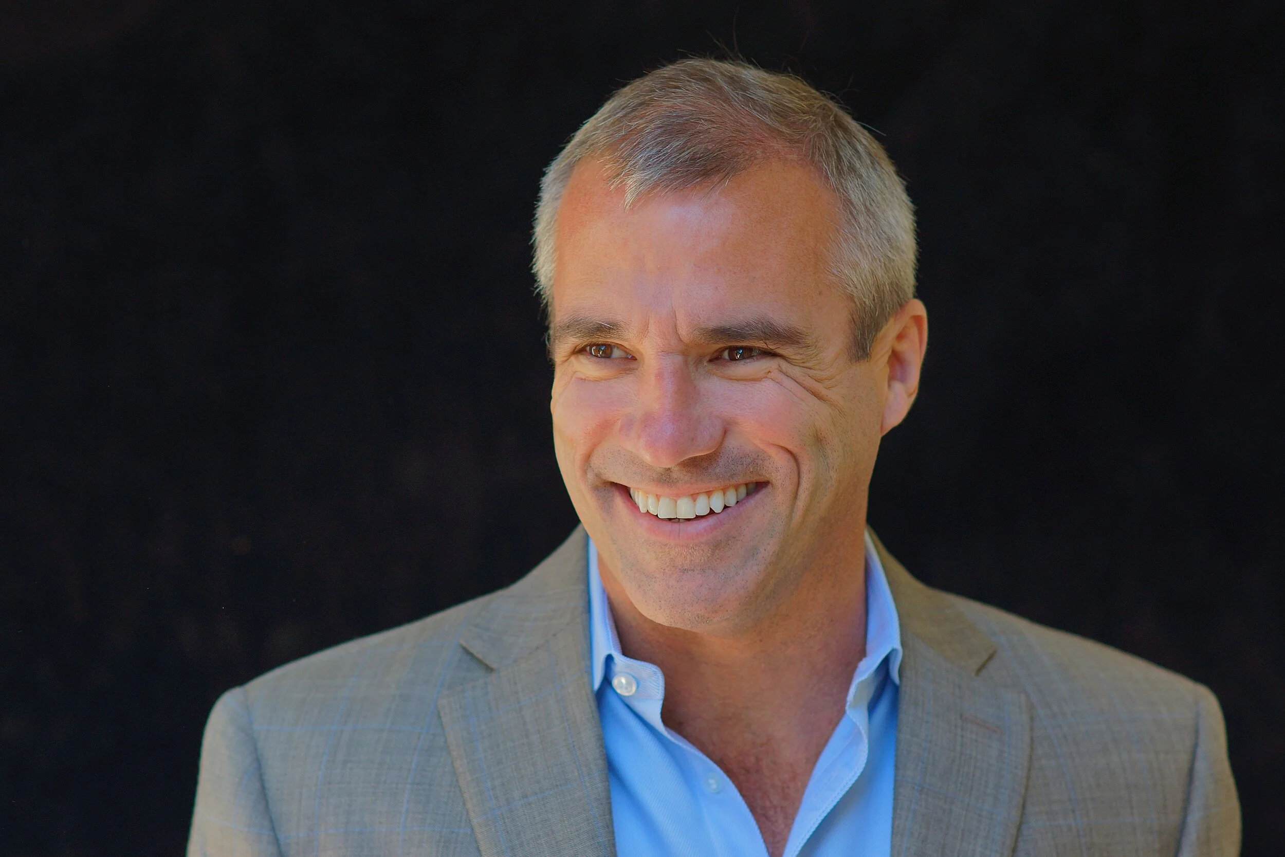 Smiling middle-aged man with short gray hair wearing a gray suit jacket and a light blue dress shirt against a dark background.