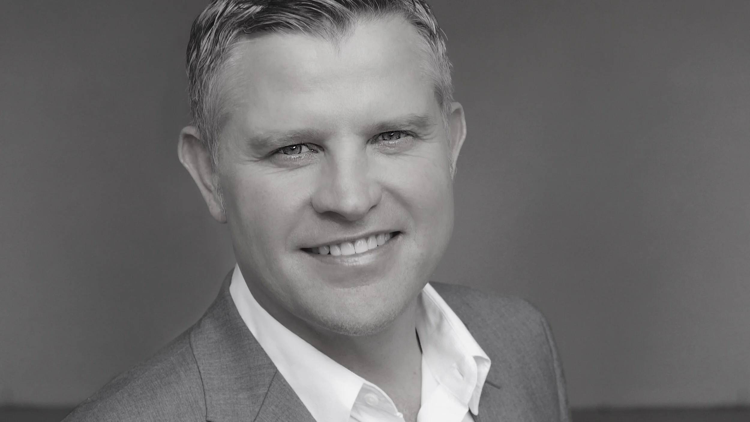 A black-and-white headshot of a smiling man with light-colored hair, wearing a suit jacket and white shirt, against a plain background.