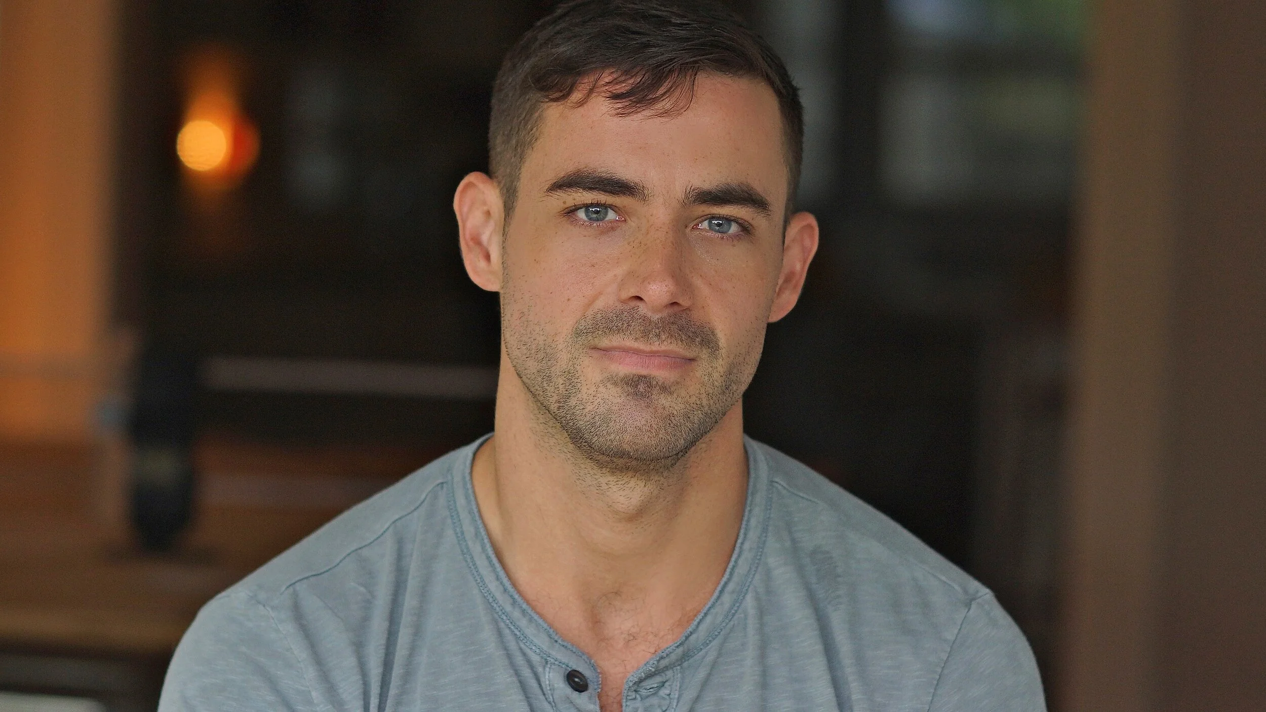 Portrait of a young man with short dark hair, blue eyes, and light stubble, wearing a gray henley shirt, sitting indoors with blurred background.