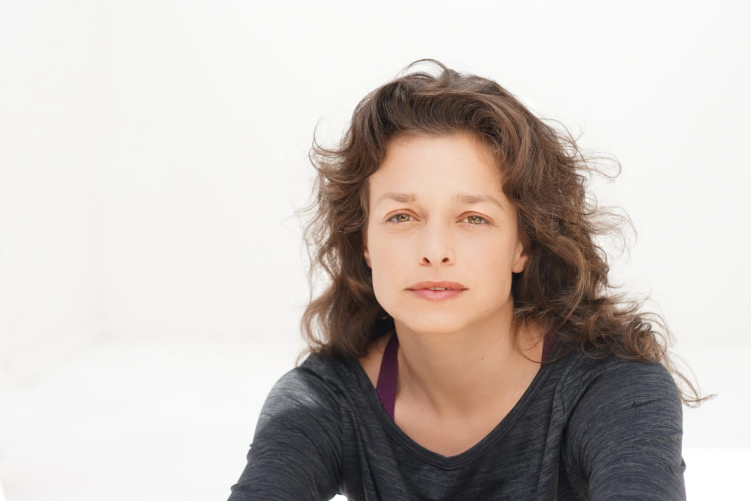 A woman with brown, curly hair and light skin, wearing a dark athletic top, looking into the camera against a white background.