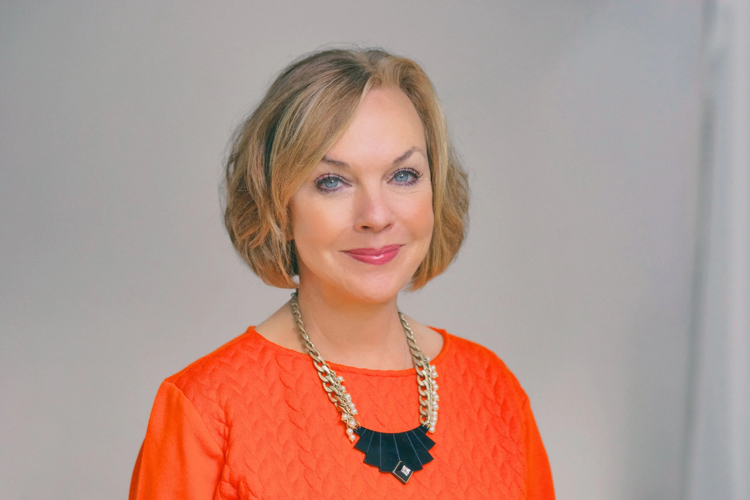 Portrait of a woman with short, wavy blonde hair wearing an orange top and a statement necklace, smiling softly against a neutral background.