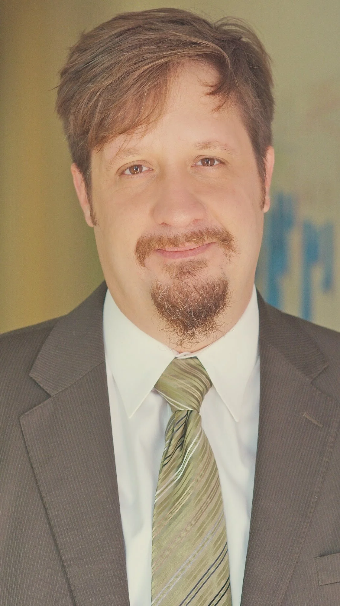 A man with brown hair and a goatee, wearing a dark suit, white shirt, and tie, smiling at the camera.