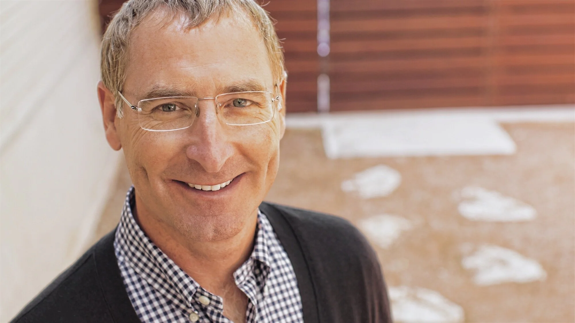A smiling middle-aged man wearing glasses, a checkered shirt, and a dark blazer, standing in an outdoor setting with a wooden fence and sand with footprints in the background.
