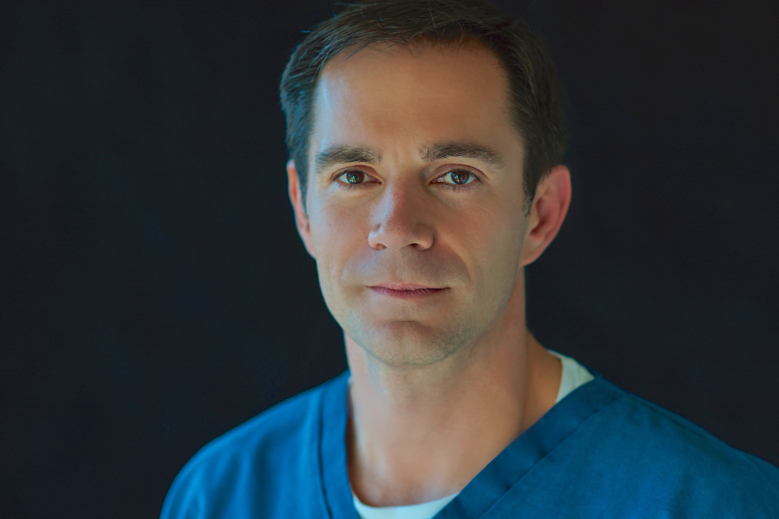 Portrait of a male medical professional wearing blue scrubs, with a neutral dark background.