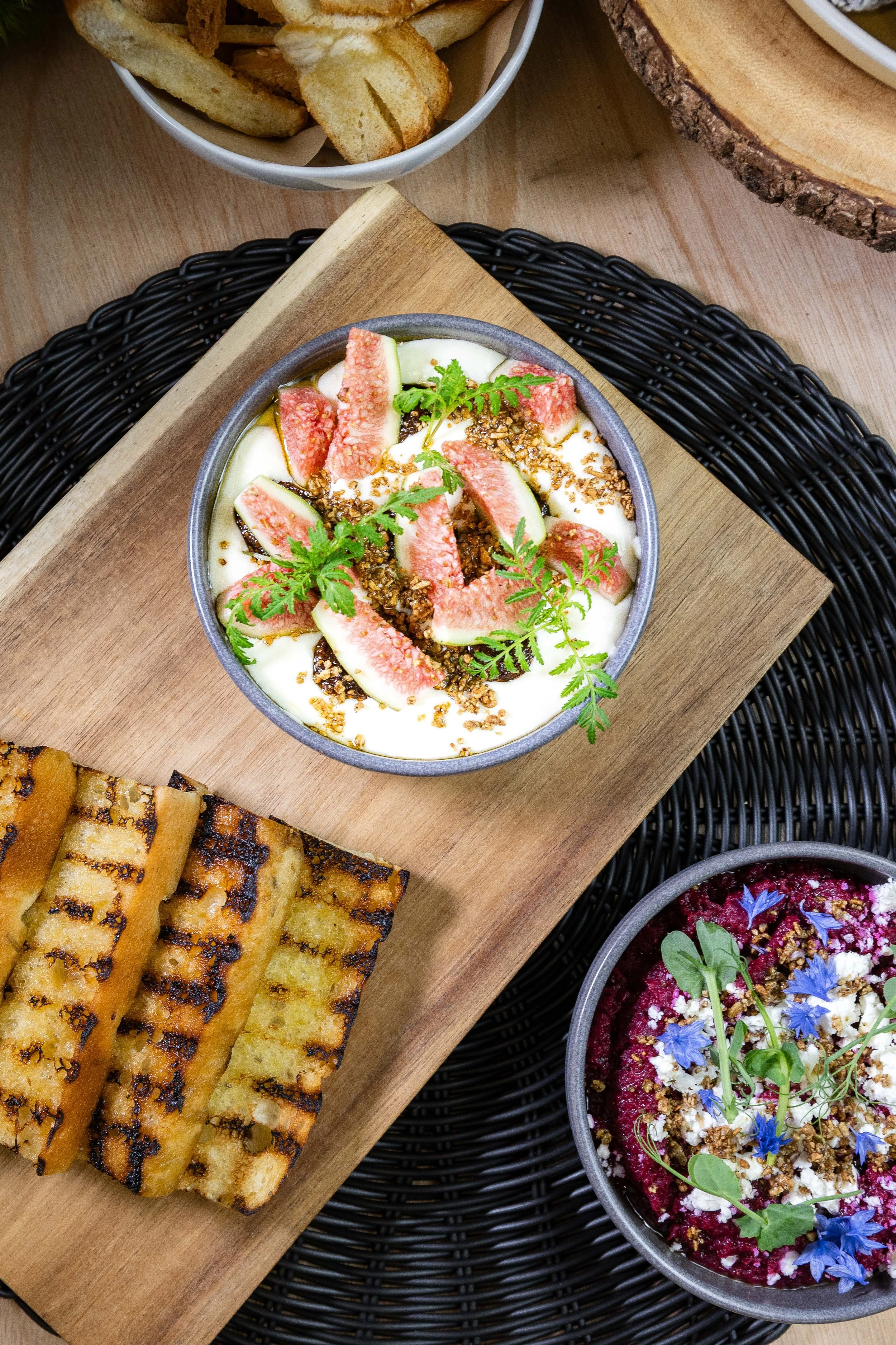 Une assiette de yaourt avec des figues fraîches, des graines et des herbes aromatiques. À côté, des tranches de pain grillé abricotés. Autres bols contenant des plats colorés, décorés avec des fleurs et des herbes.
