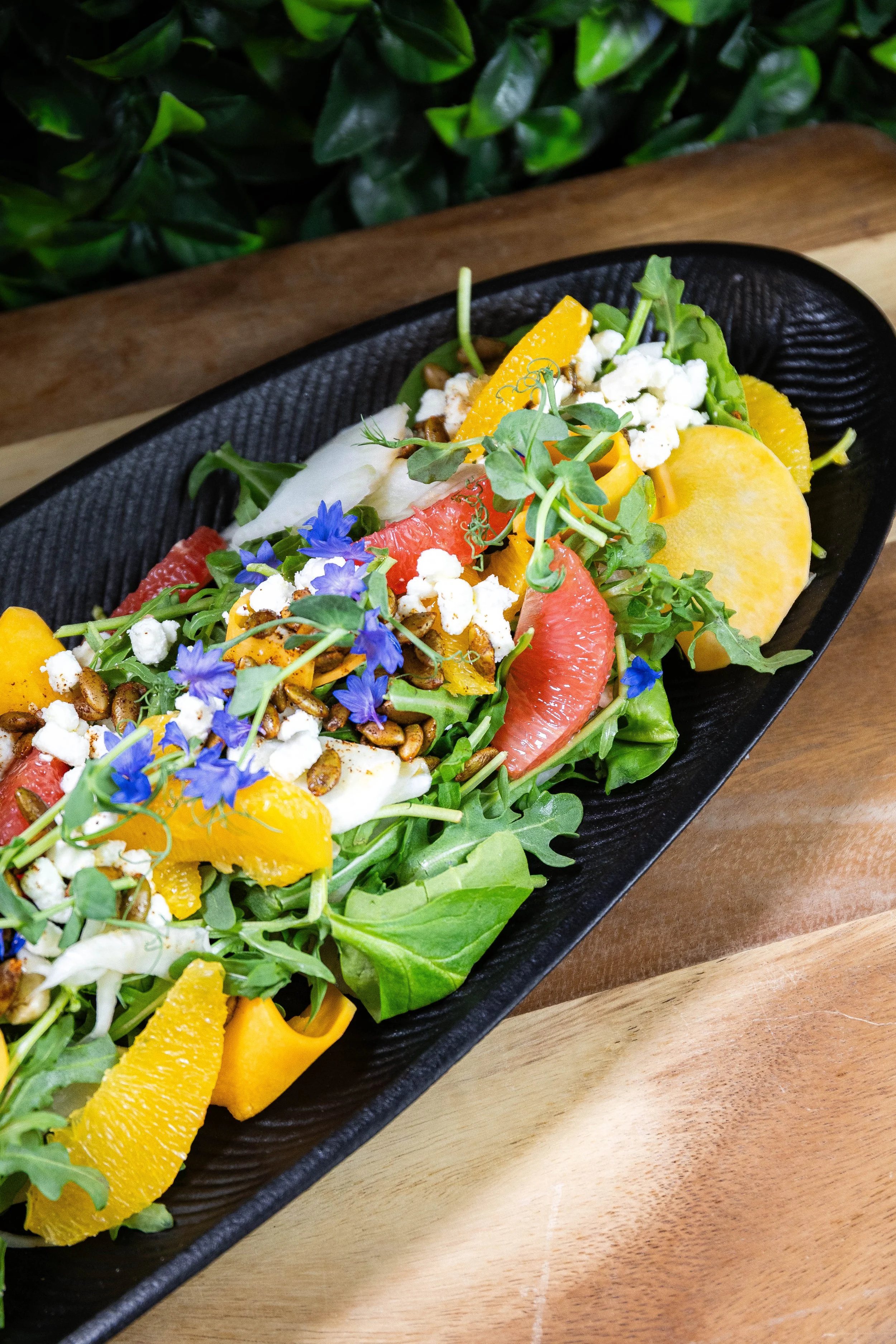 Salade colorée avec des feuilles vertes, des segments de pamplemousse, des tranches de poivron jaune, de la feta, des graines et des fleurs violettes dans un plat noir.