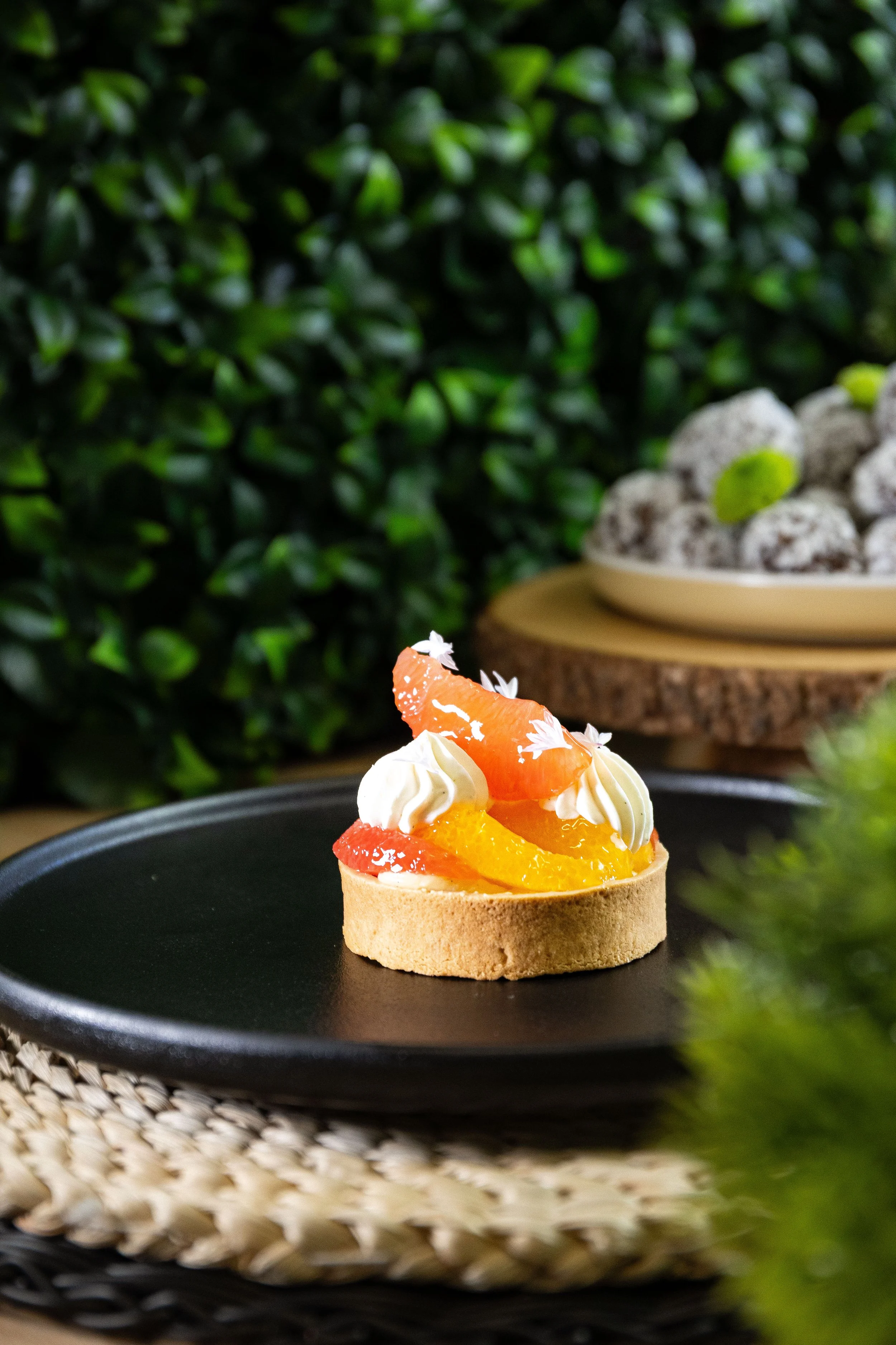 Tartelettes aux fruits avec crème fouettée, oranges et citrons, présentées sur une assiette noire devant un fond de buissons verts et un plateau avec des biscuits en arrière-plan.