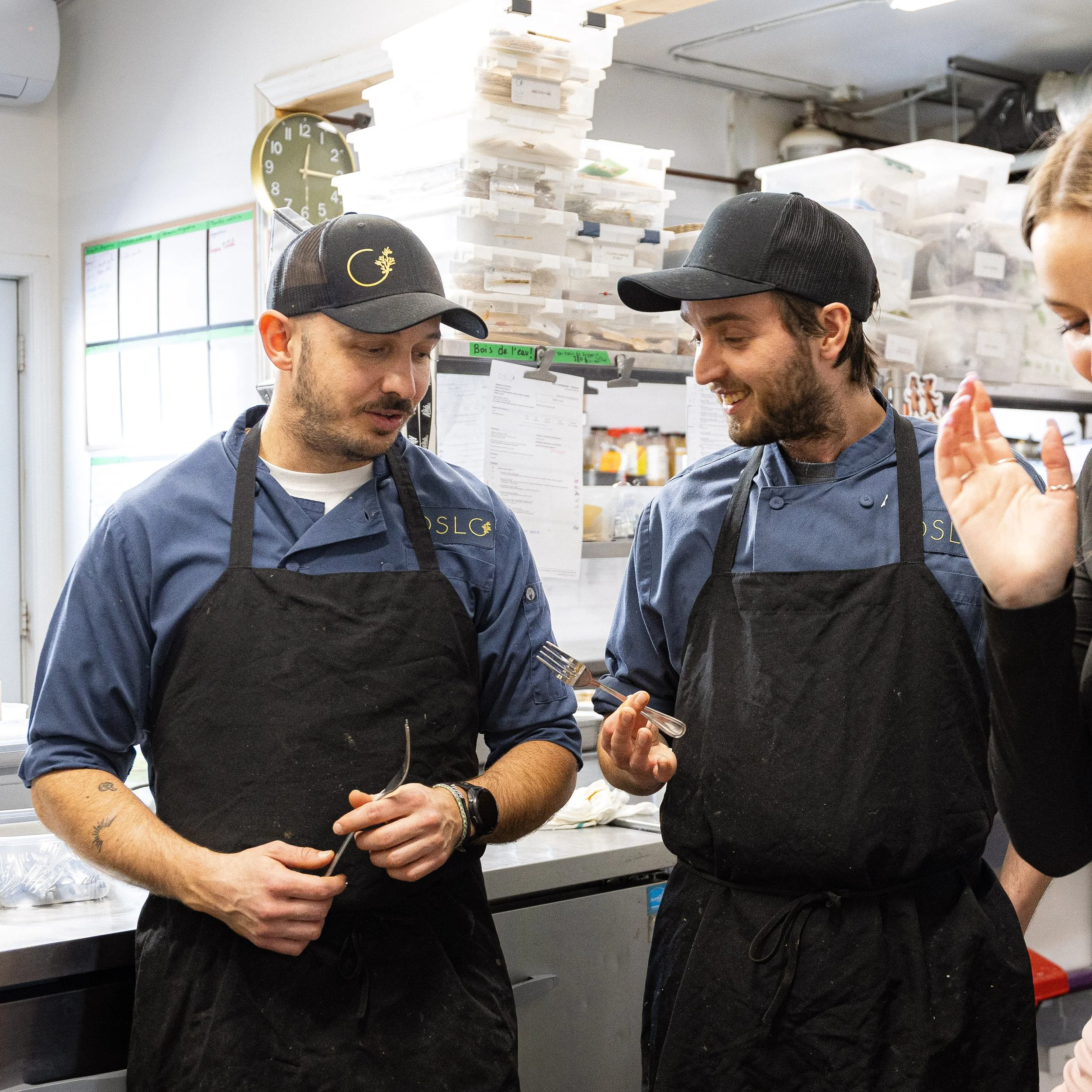 Deux hommes en uniformes de cuisine discutent dans une cuisine professionnelle, portant des tabliers noirs et des casquettes noires.