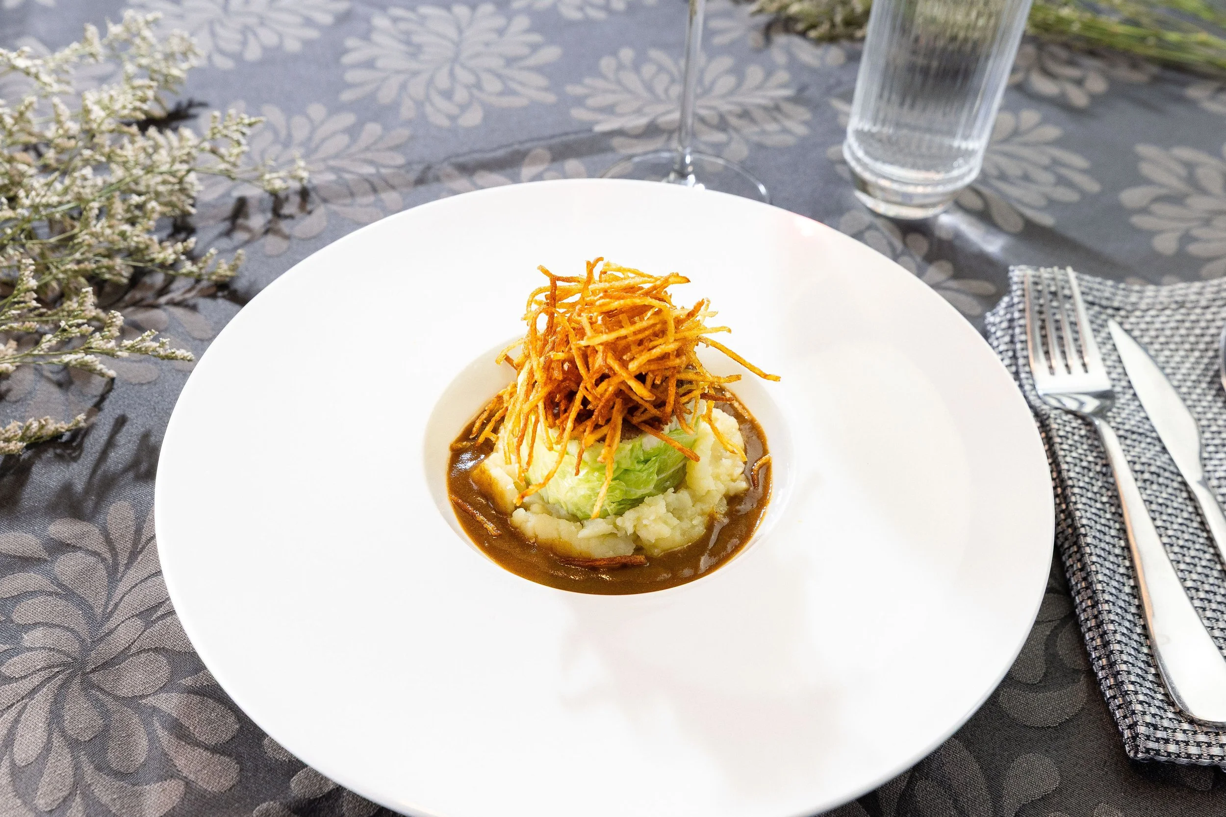 Assiette avec viande, légumes, microgreens, et sauce, accompagnée d’un verre de vin rouge et d’un verre d’eau sur une table en plein air.