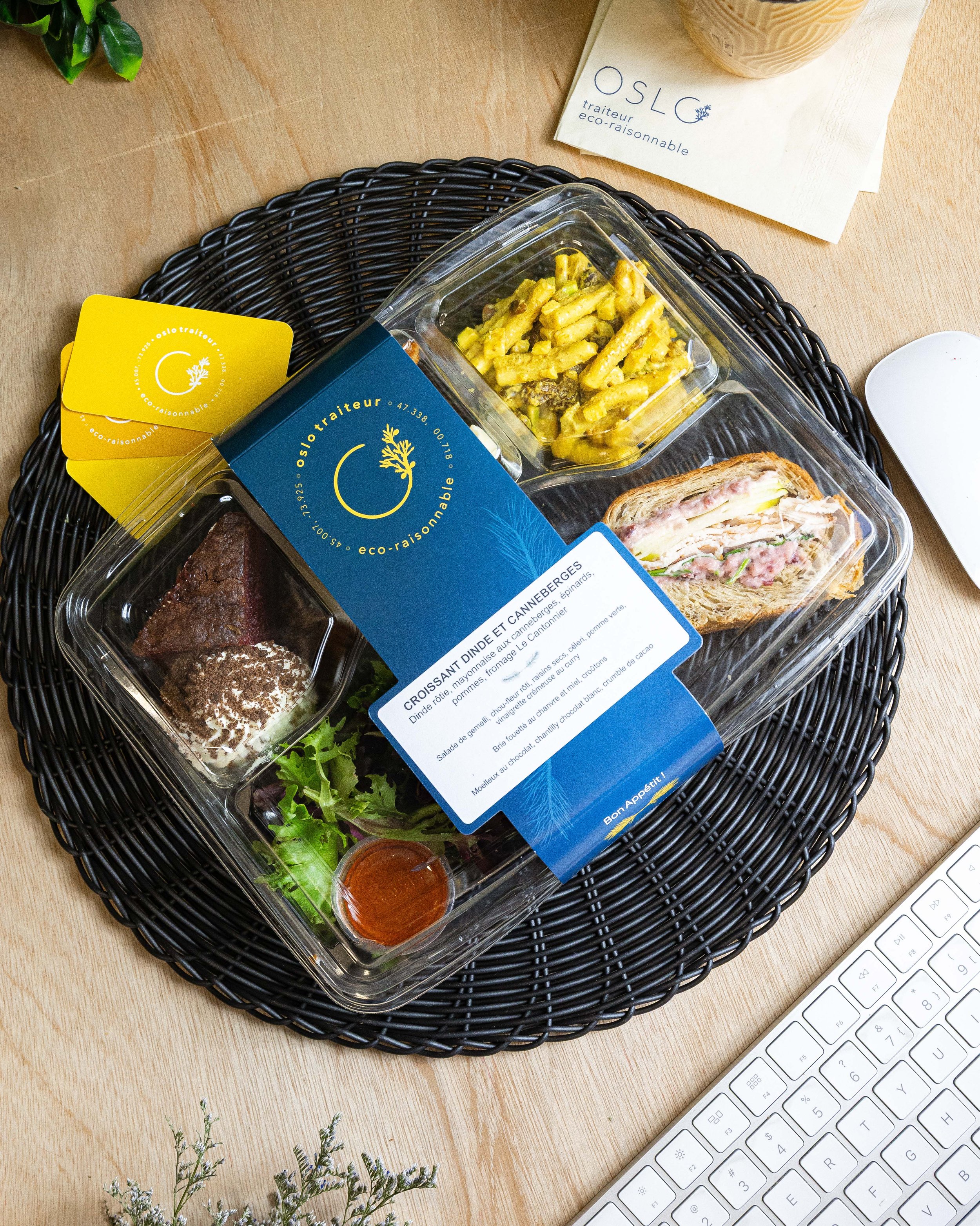 Une boîte repas contenant un sandwich, une salade, et un morceau de gâteau, placée sur un plateau en rotin noir, avec des cartes de visite jaunes et un sac en papier blanc en arrière-plan.