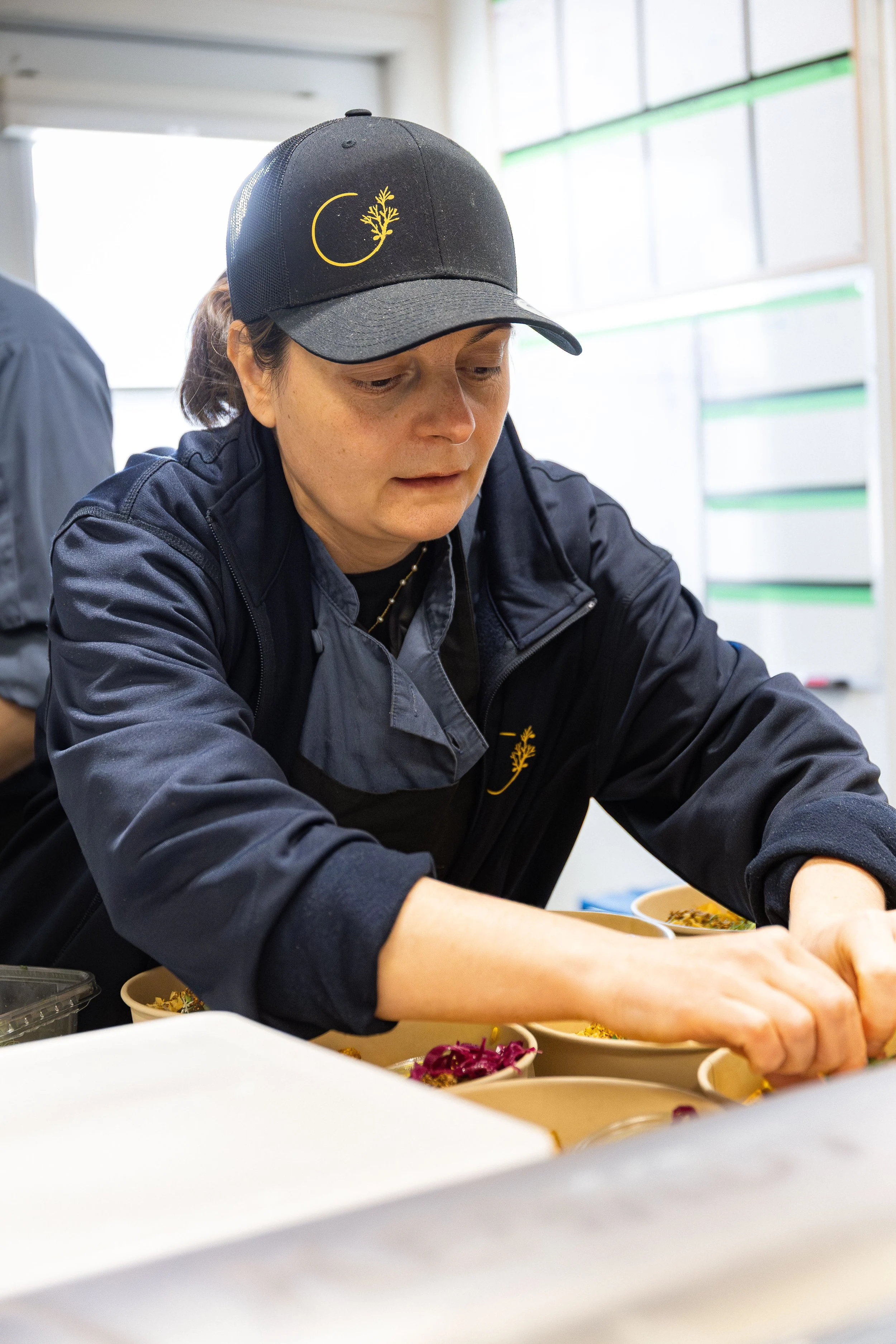 Une femme portant une casquette noire et un uniforme noir prépare des plats dans une cuisine.