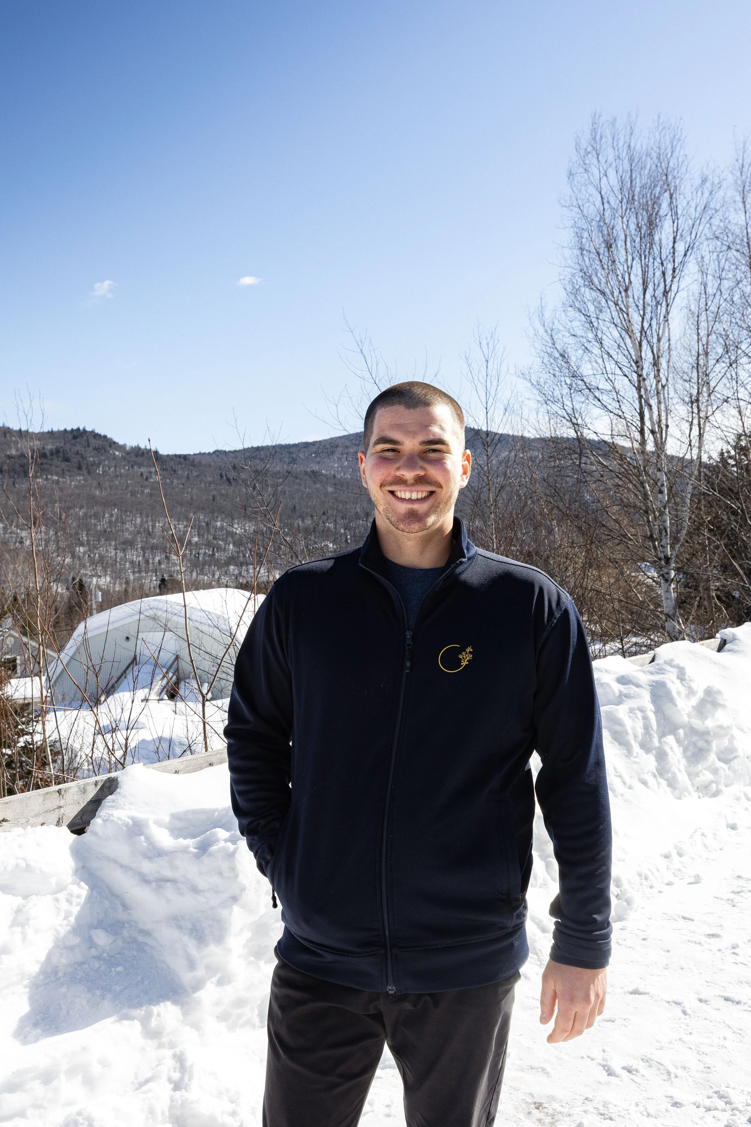 Un homme souriant debout dans la neige, portant une veste noire, en arrière-plan des montagnes et des arbres nus sous un ciel bleu clair.