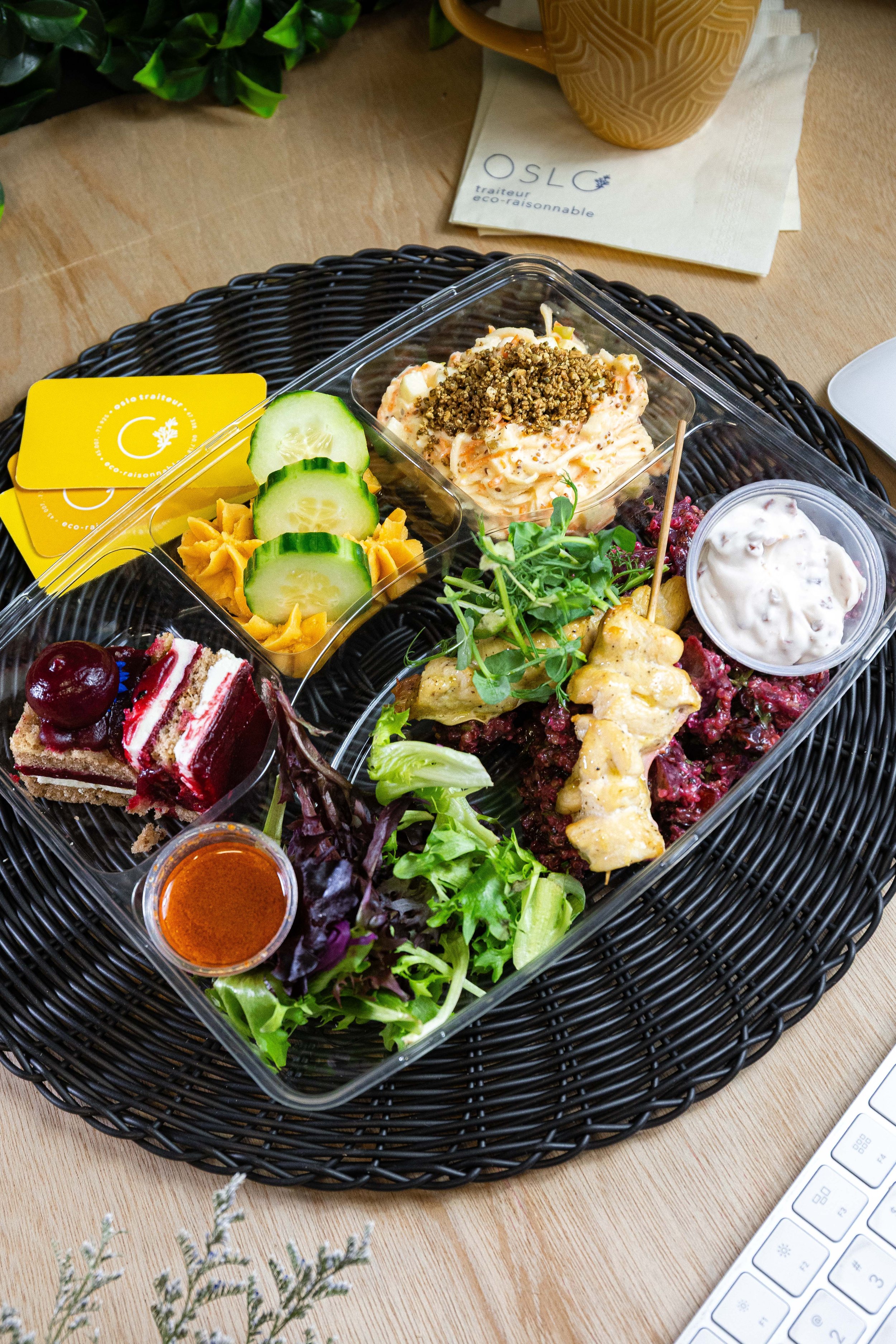 Assiette de plats végétariens ou vegans comprenant une salade verte avec des pousses, du borscht aux betteraves, un tartare végétal ou de poisson, une petite portion de gâteau ou entremets, des rondelles de concombre, une sauce ou vinaigrette, et une cuillère de sauce ou de yaourt, présentés dans des récipients en plastique sur un plateau noir.
