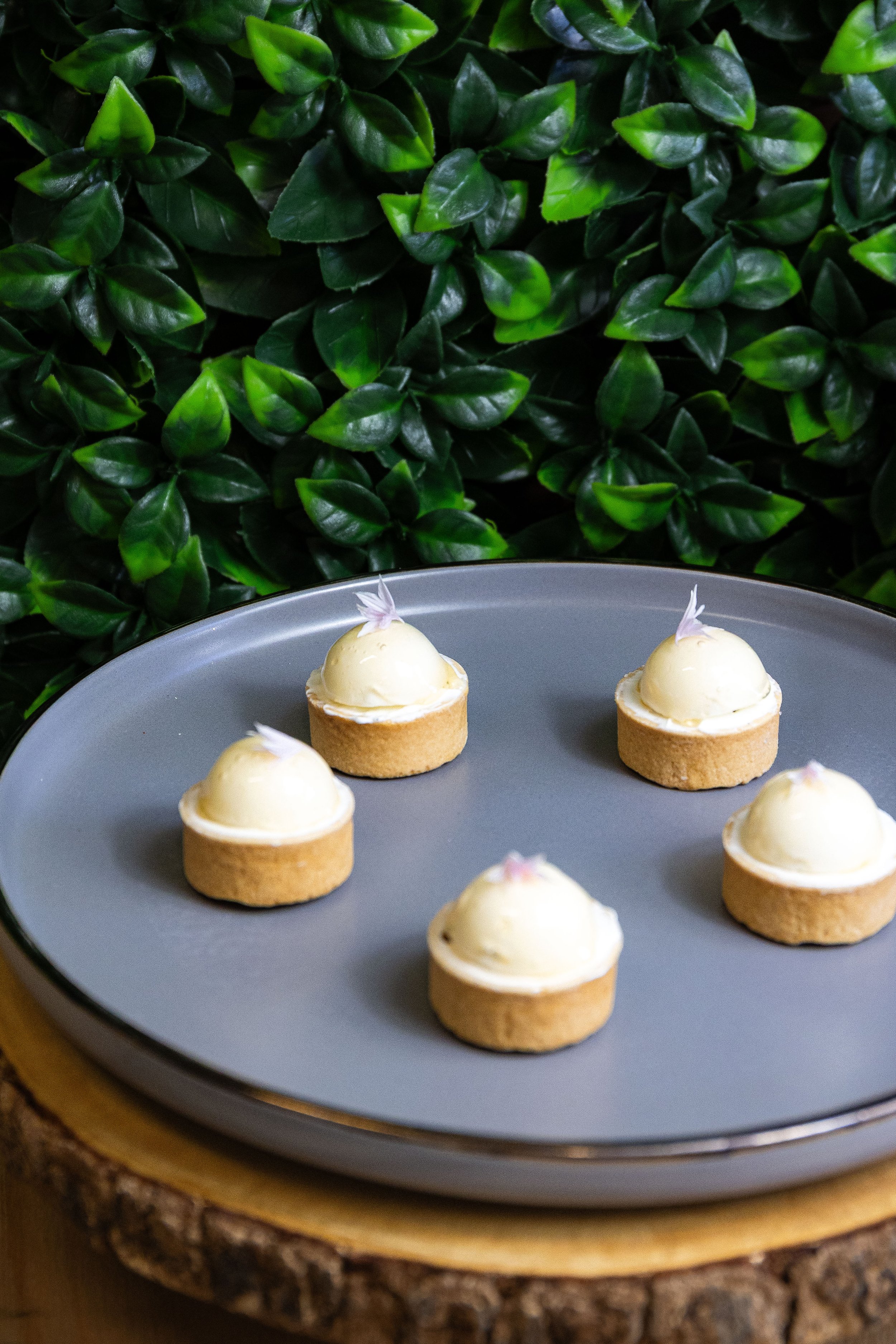 Petits entremets à la vanille avec une tête blanche, disposés sur un plateau gris, avec un décor de feuilles vertes en arrière-plan.