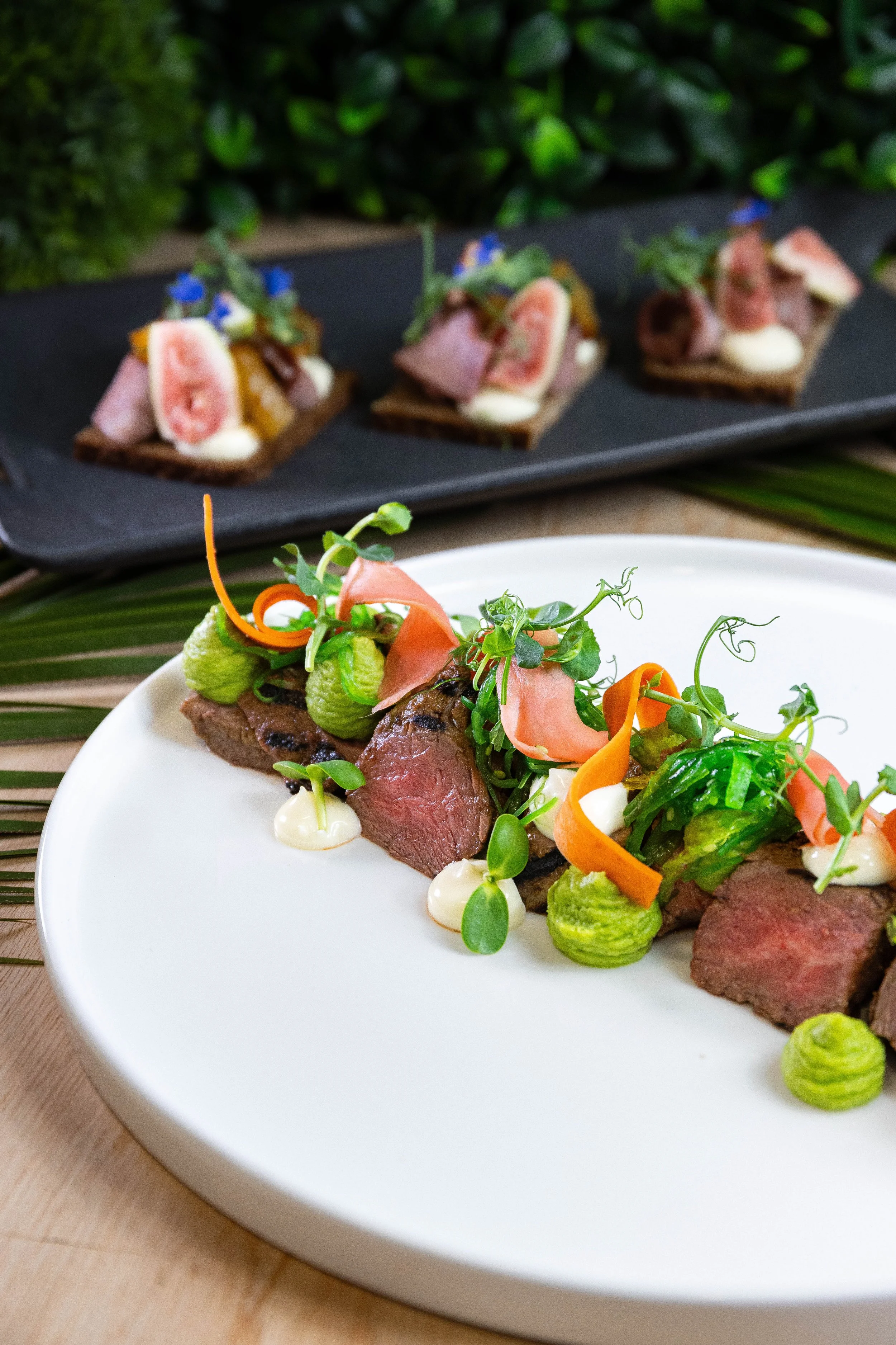 Assiette de viande rouge avec légumes garnis de microgreens, accompagnée de sauces blanches, en premier plan. En arrière-plan, des petits doigts de viande avec garnitures végétales sur une plaque noire.