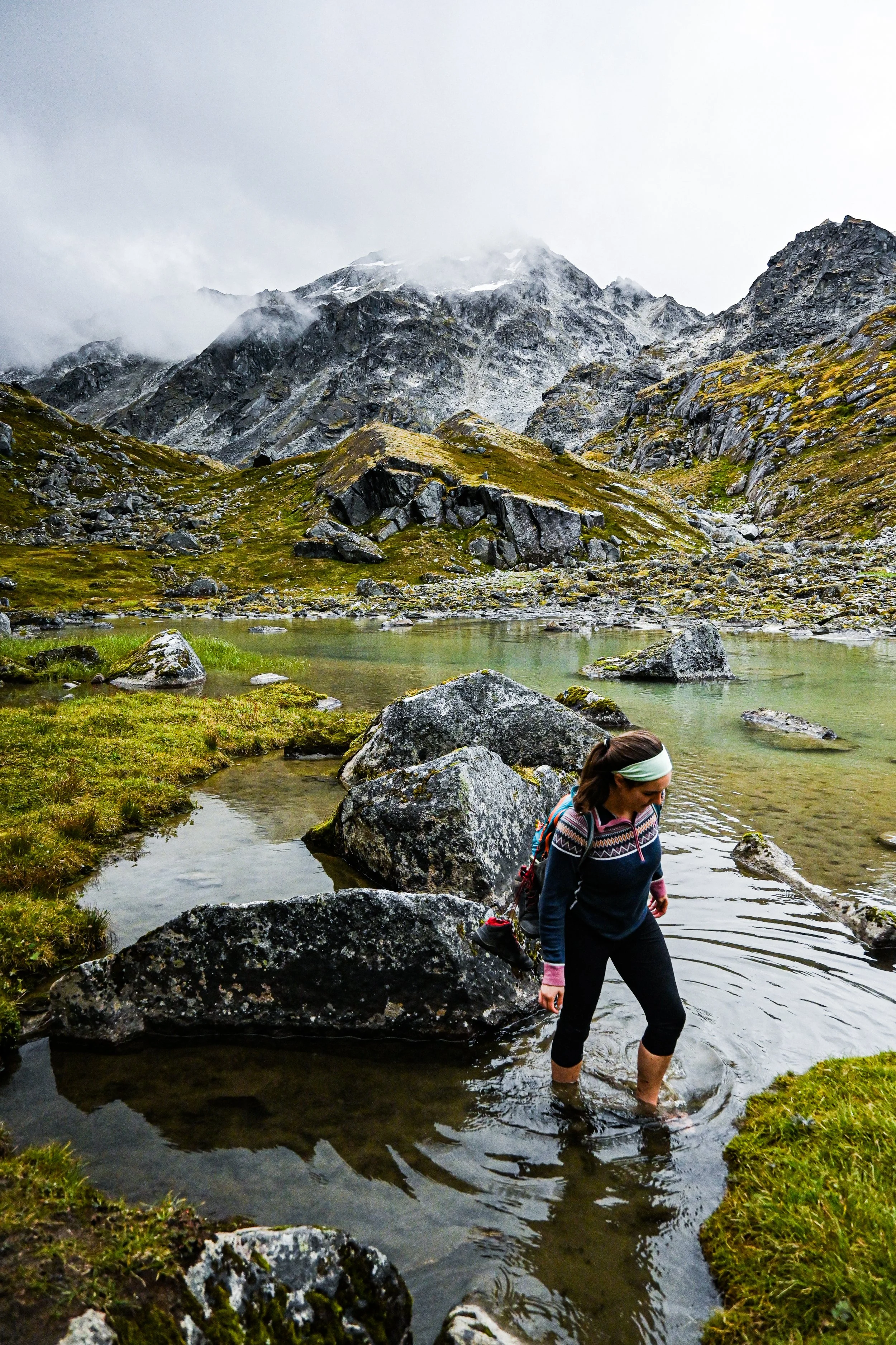 A person crossing a small stream in a mountainous landscape with rocky hills and snow-capped peaks, cloudy sky.