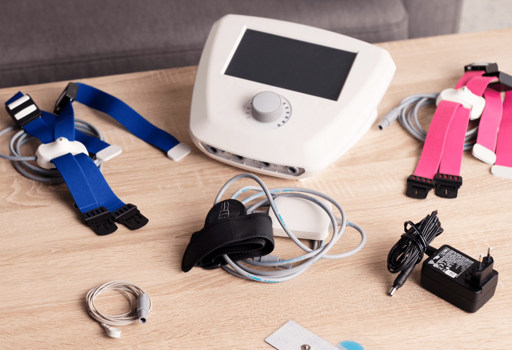 A collection of medical devices on a wooden table, including a white monitor, colorful straps with sensors, a blood pressure cuff, a power adapter, and various cables.