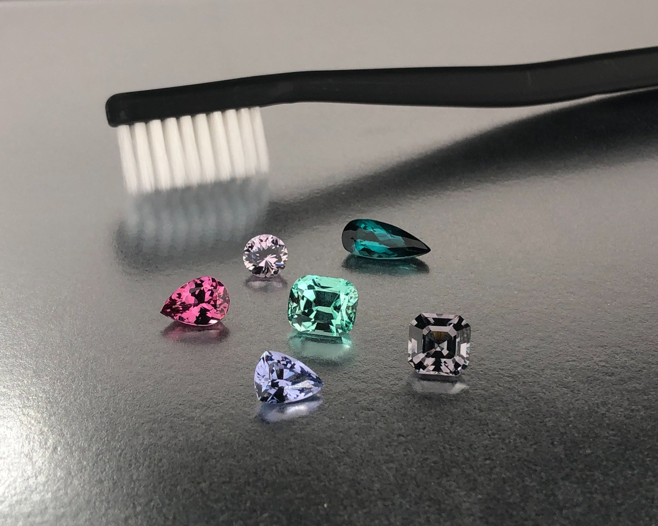A set of seven multicolored gemstones arranged on a gray surface with a black toothbrush near the top left corner.