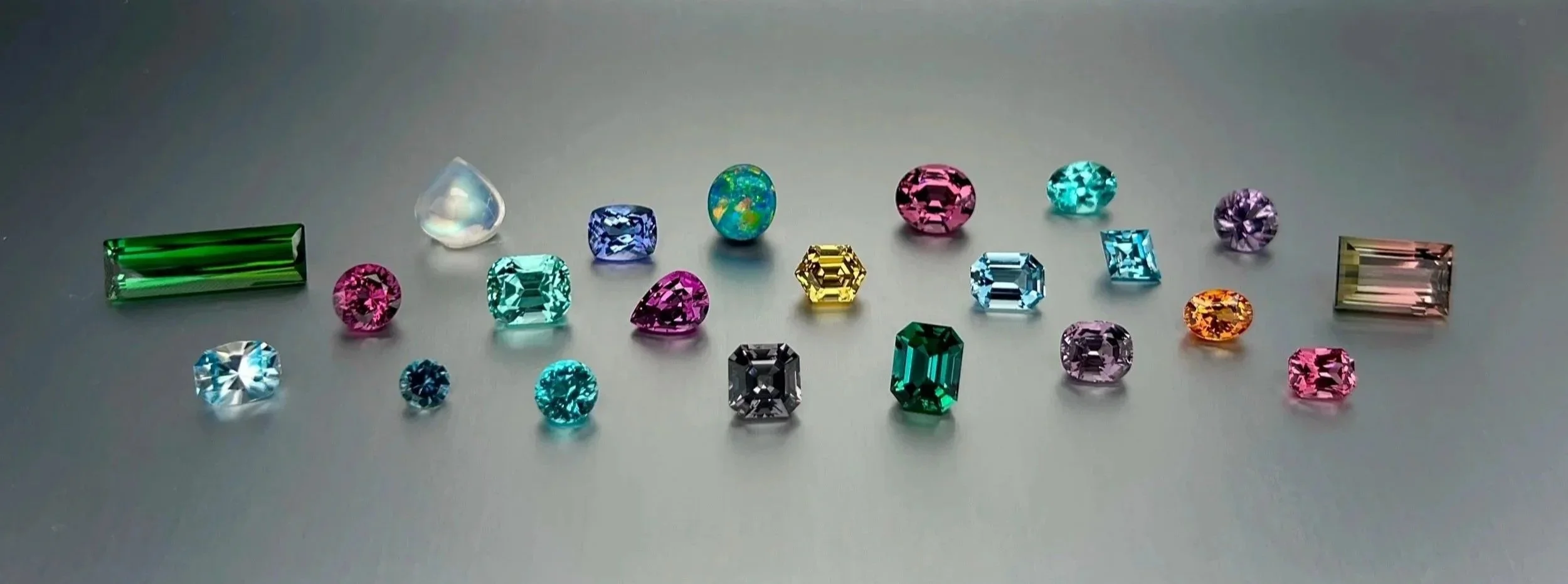 Collection of natural colored gemstones including tourmaline, sapphire, spinel, zircon, garnet, rainbow moonstone, opal, and chrysoberyl available for custom jewelry design and gemstone sourcing.