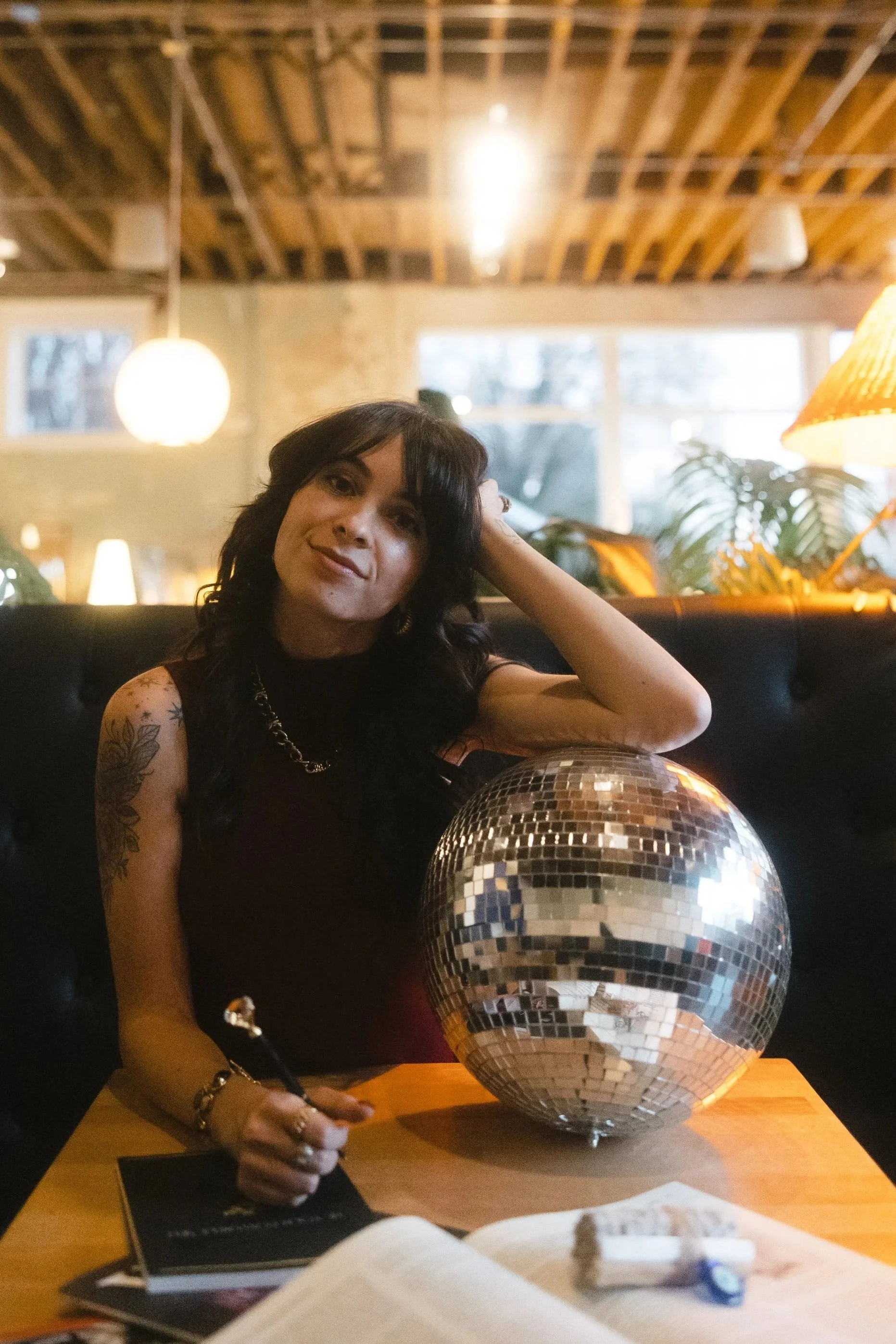 A woman with dark hair and tattoos on her arm sitting at a table in a cozy, warmly lit cafe or bar, holding a pen, with a disco ball on the table and a notebook in front of her.