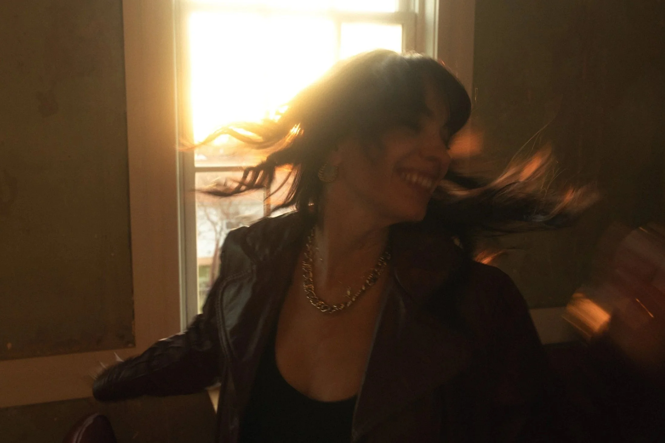 A woman with dark hair smiling and looking to her left, illuminated by sunlight streaming in through a window behind her.