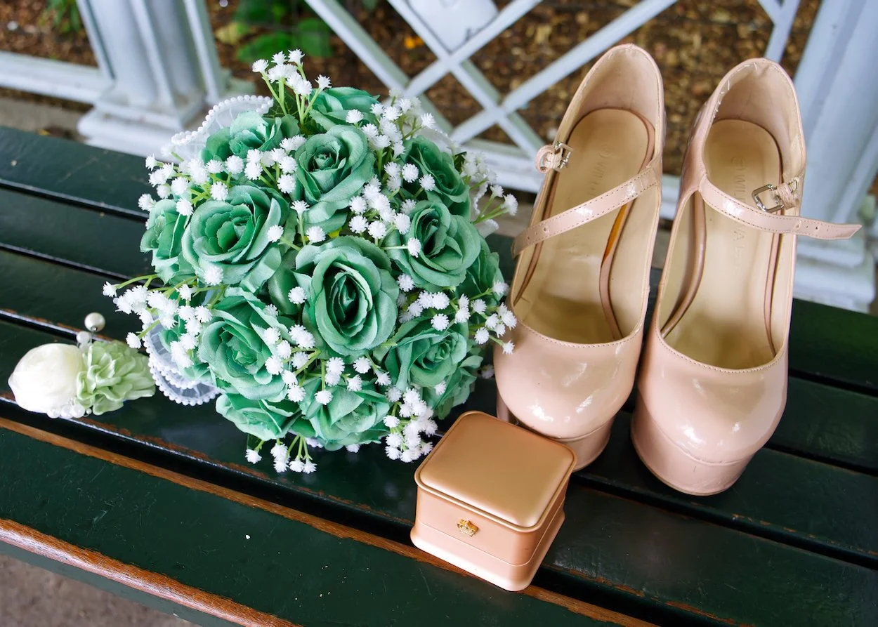 Wedding Shoot: A pair of beige high-heeled shoes, a bouquet of green roses with white accents, a small gold jewelry box, and a white rose boutonniere arranged on a green wooden bench.