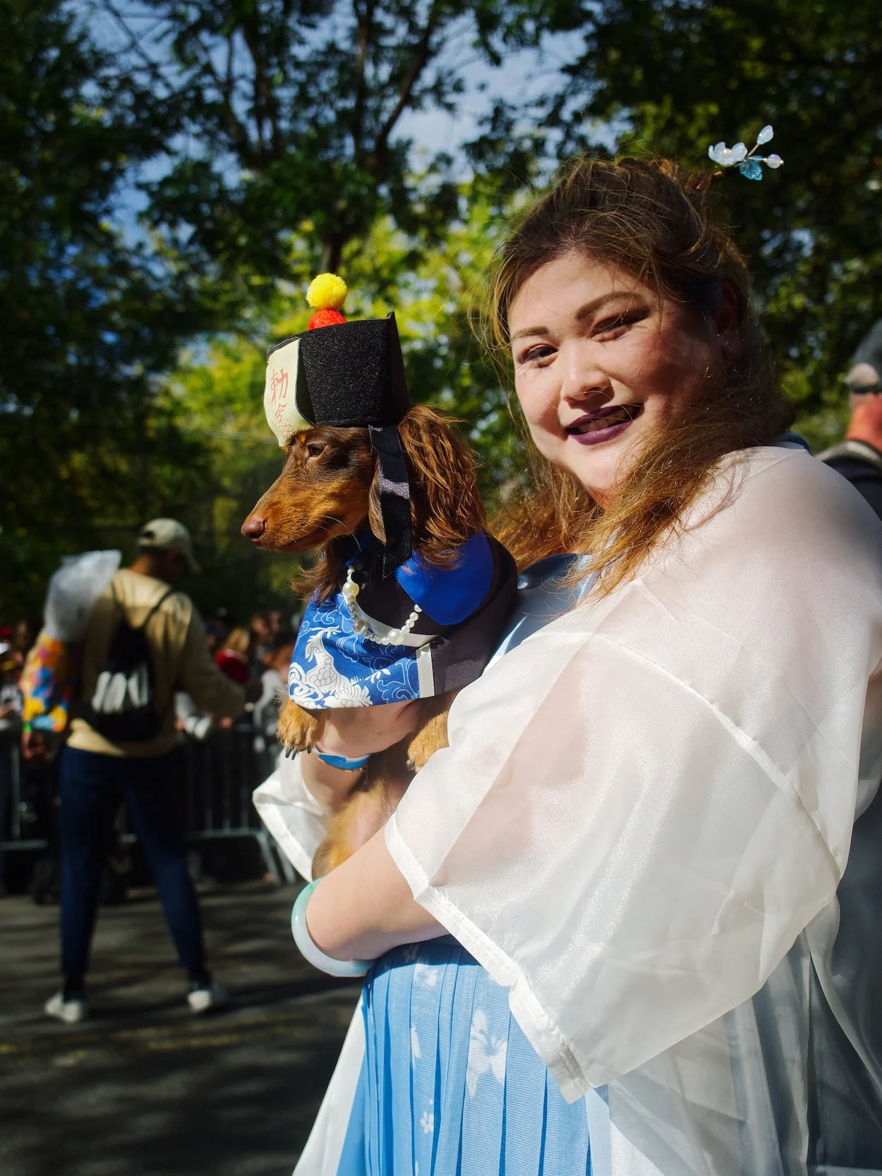 Event Portrait, Halloween Dog Parade: Woman with light skin and dark lipstick holding a Dachshund dressed in traditional Japanese clothing and a hat, at an outdoor event with trees and other people in the background.
