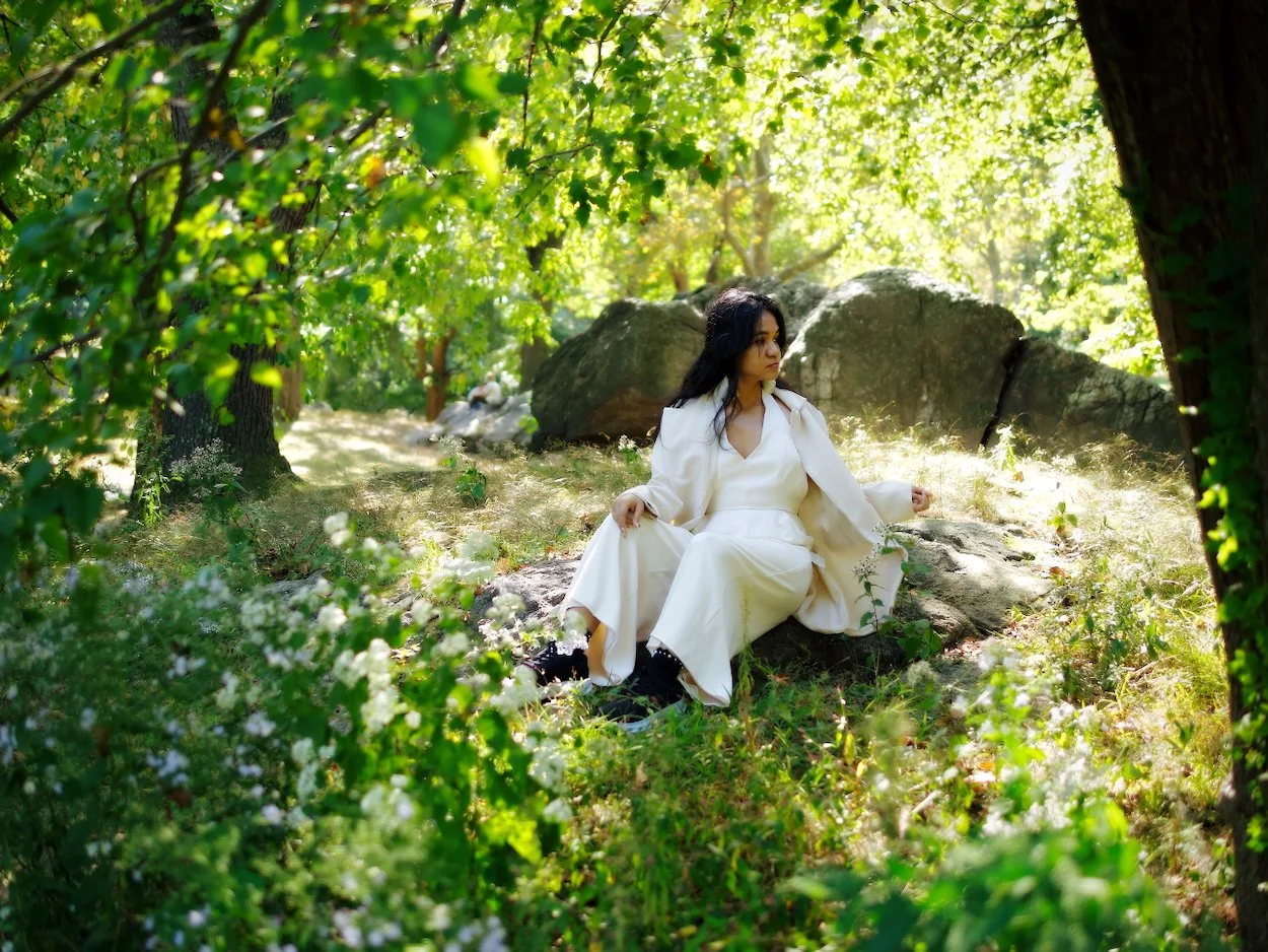 Central Park Portrait: A woman in white clothing sitting on a rock in a lush green forest, surrounded by trees and sunlight.